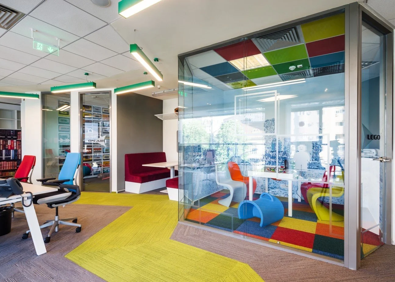 Modern office space with colorful furniture, glass enclosed playroom with LEGO building set, bright lighting, and large windows.