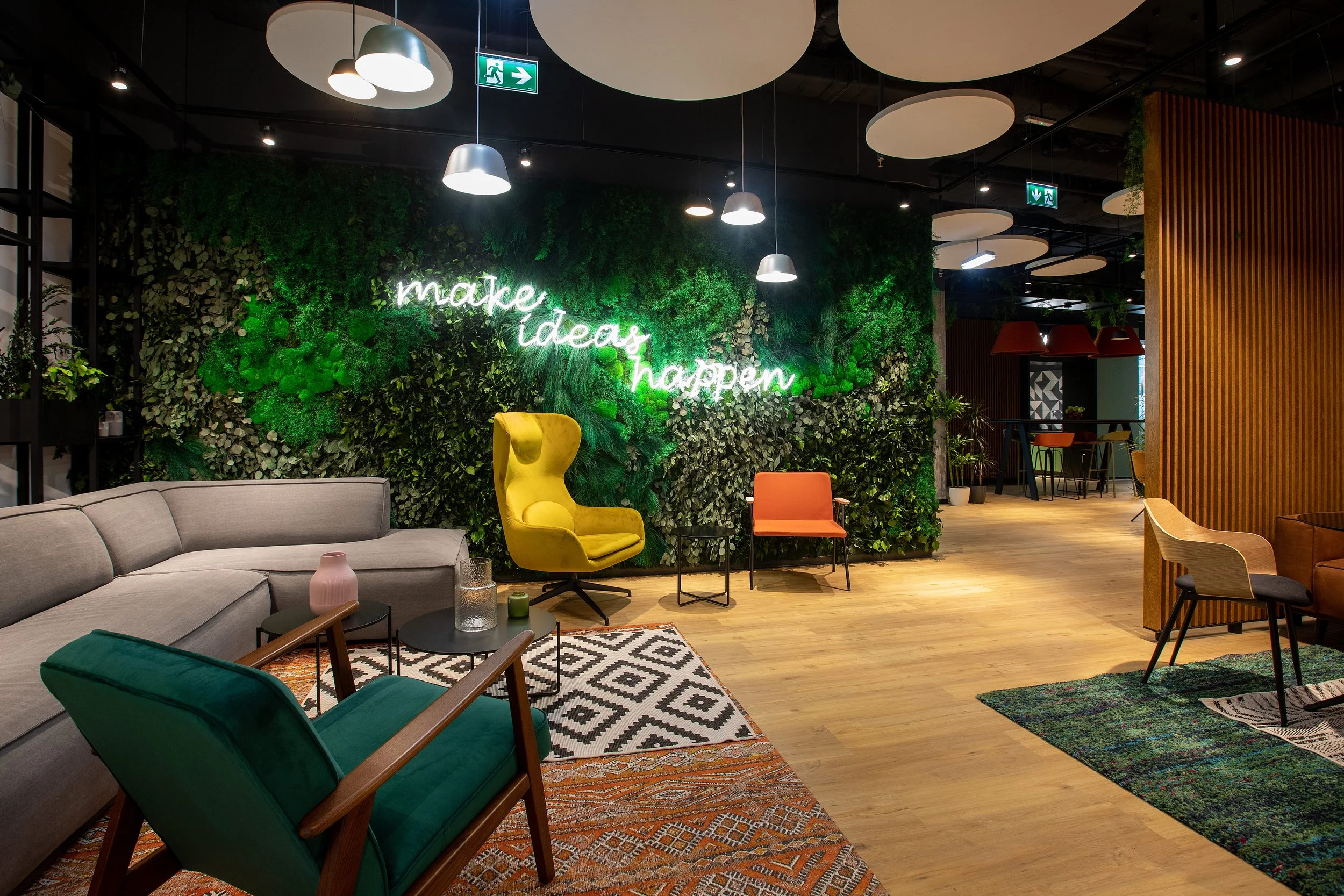 Interior of a modern lounge with a green wall decorated with plants and a neon sign saying 'make ideas happen'. The space features a beige sectional sofa, a green armchair, a yellow high-backed chair, and various small tables, with wooden flooring and ceiling lights.