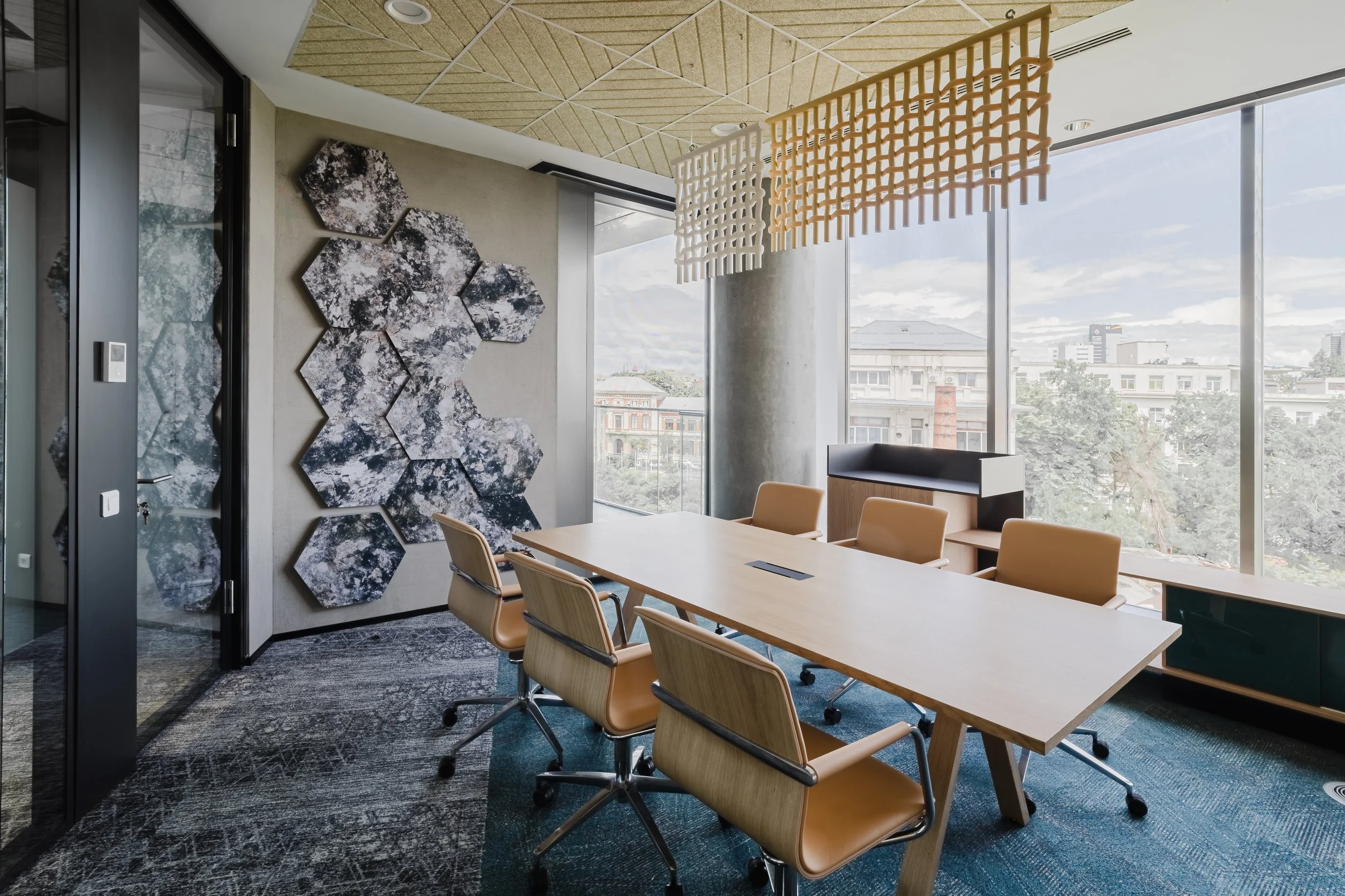 Modern conference room with large window, wooden table surrounded by six beige chairs, decorative wall art with hexagonal panels, and abstract fabric ceiling art.