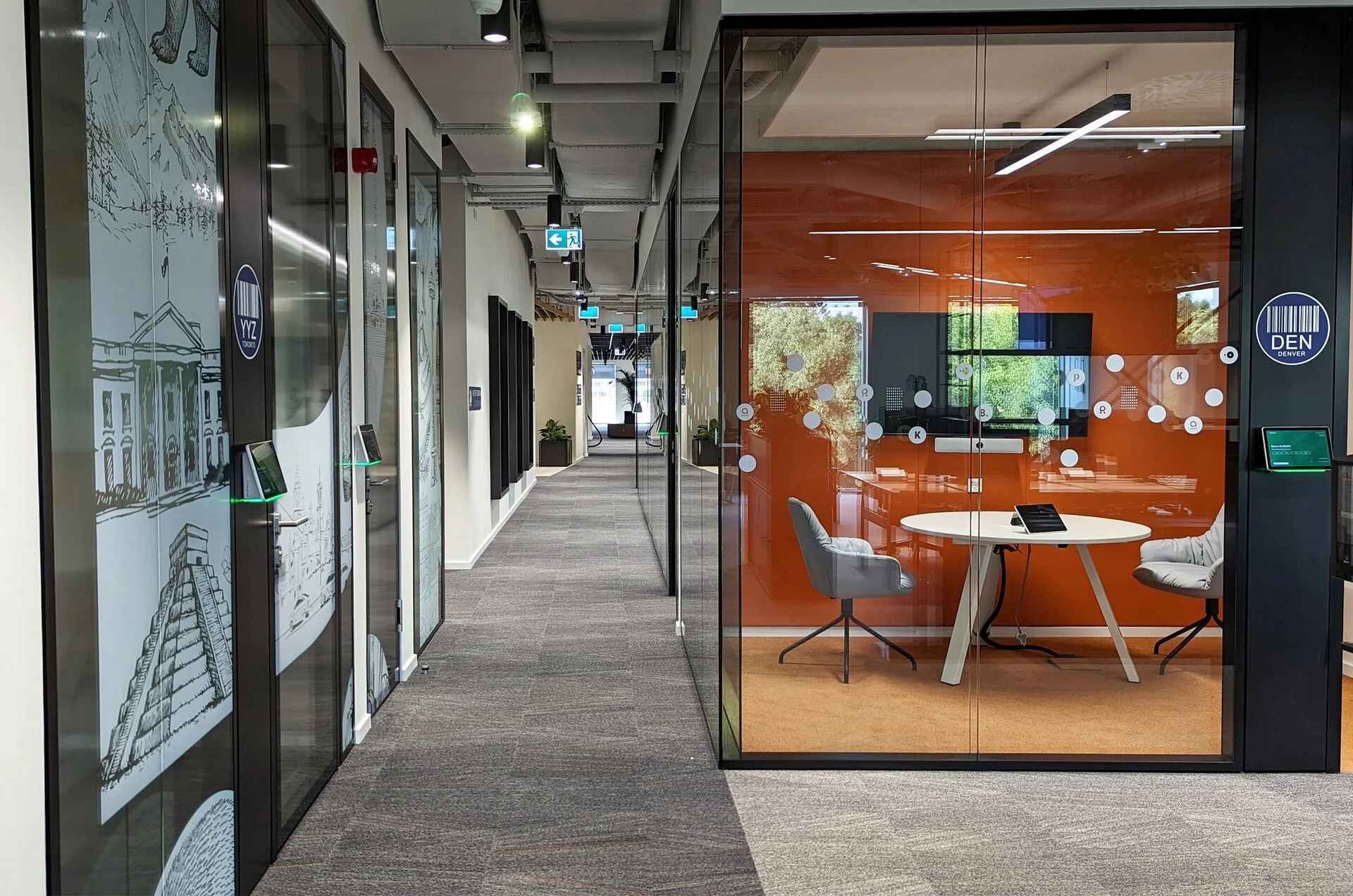 Modern office hallway with glass conference room on the right, orange accent wall, table and chairs inside, hallway has patterned carpet, wall art, and ceiling lighting fixtures.