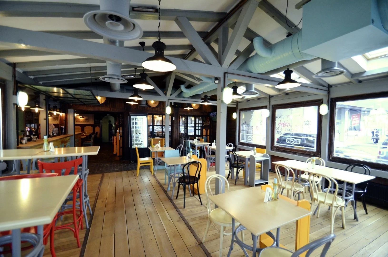 Empty cafe with multiple colorful chairs and tables, large windows, and industrial ceiling with exposed ductwork and lights.