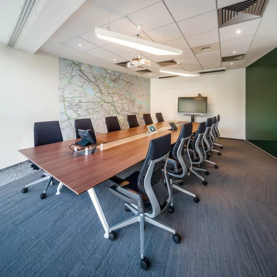 Empty conference room with a long wooden table, black office chairs, a large map on the wall, a wall-mounted TV, and a ceiling with lights and air vents.