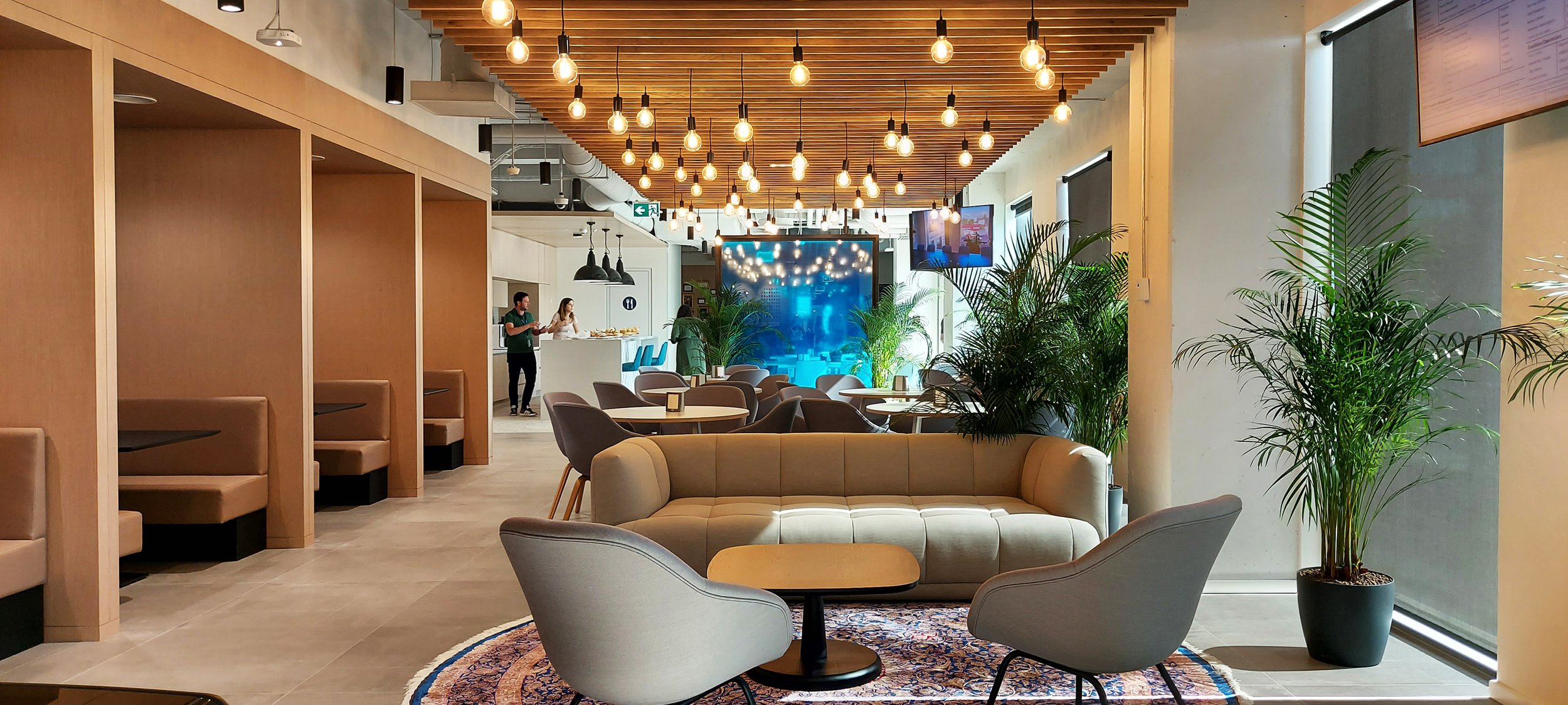 Modern lounge area with beige sofa, two armchairs, and potted plants. Ceiling decorated with hanging Edison bulbs. There are tables, a barista serving coffee, and screens on the walls.
