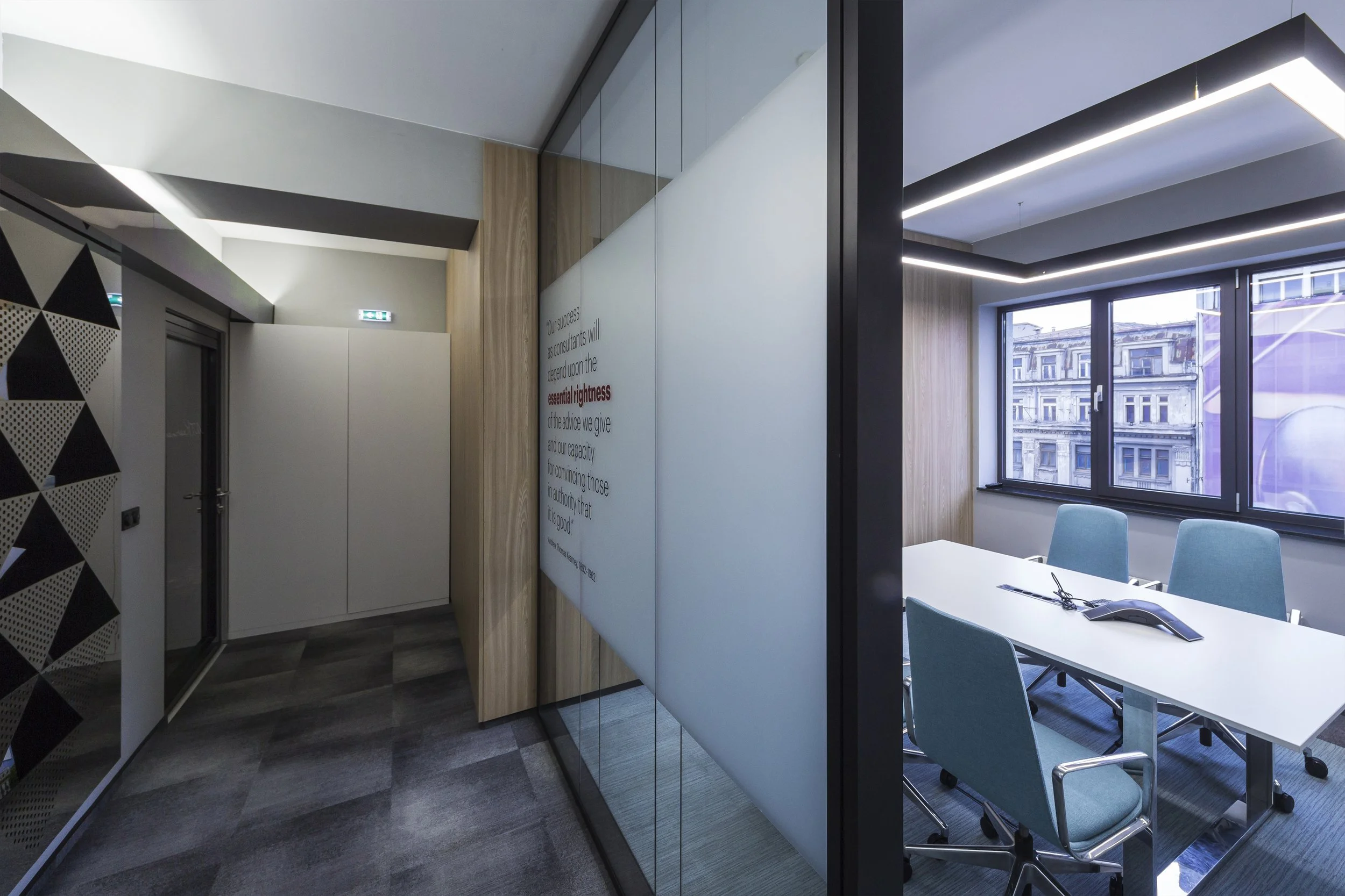 Modern office conference room with large window, blue chairs around a white table, and a built-in ceiling lighting fixture.