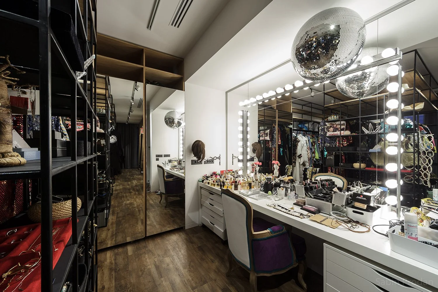 A dressing room or makeup area with a large mirror surrounded by lights, a vanity table cluttered with cosmetics, jewelry, and accessories, a chair, and shelving filled with clothing and accessories, with disco balls hanging from the ceiling.