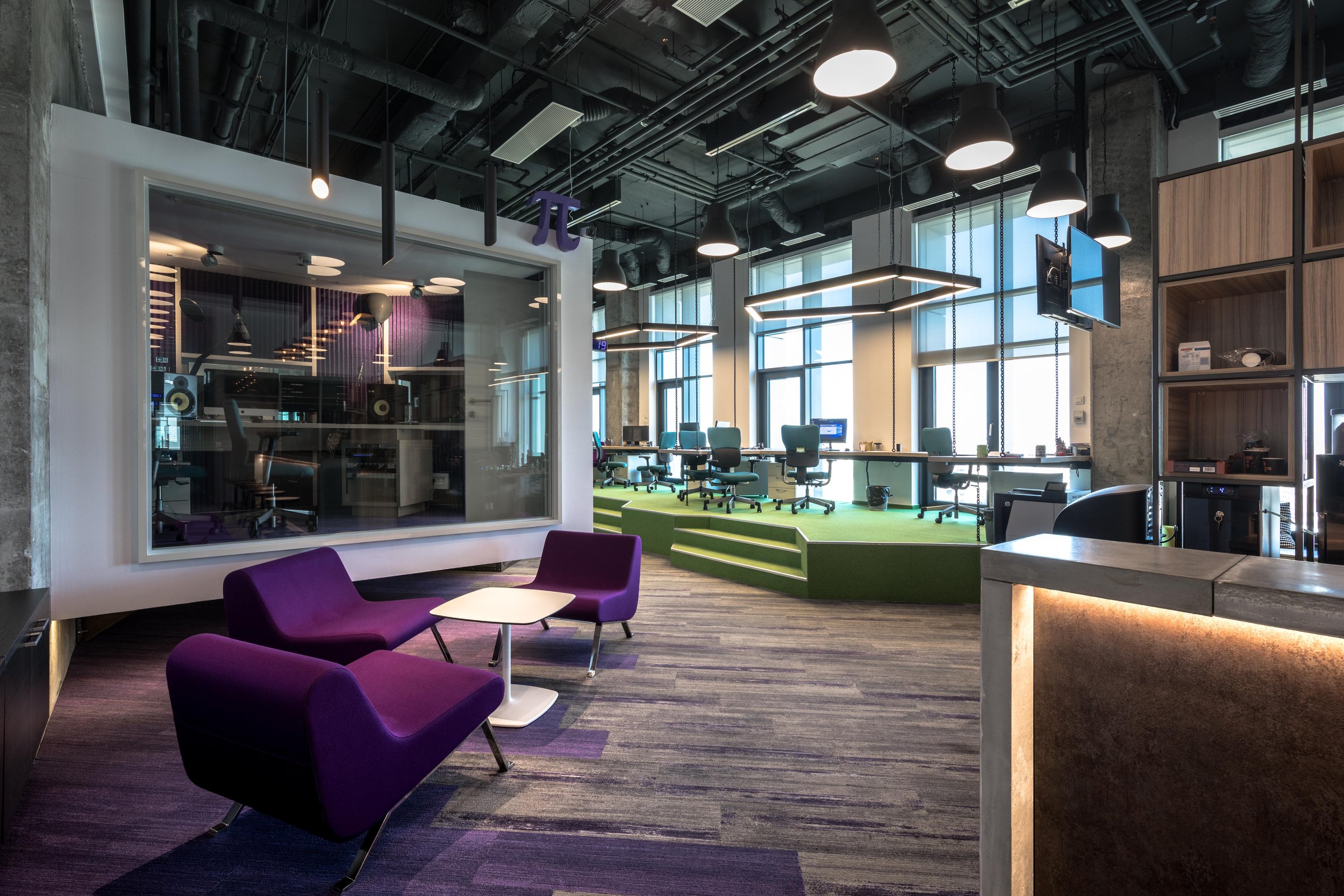 Modern office space featuring a music recording studio with soundproof glass, a seating area with purple chairs and a white table, high workstations with green carpeting, and large windows allowing natural light.