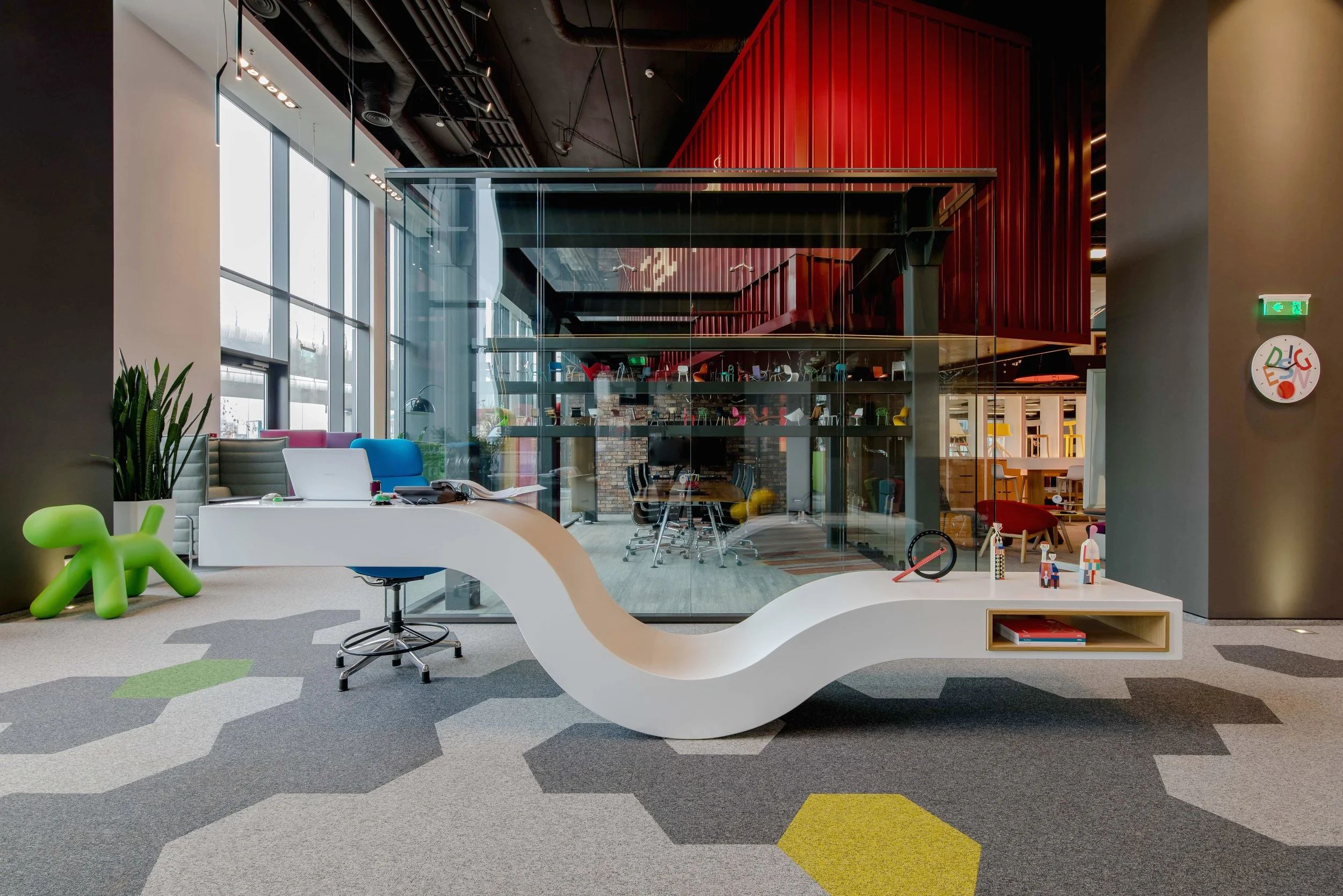 Modern office space with a white, curvy reception desk, colorful chairs, a glass-walled conference room, and playful decor including a green dog-shaped sculpture and various toys.
