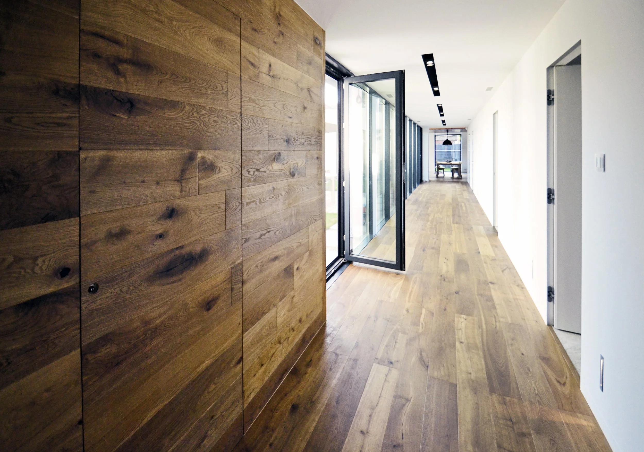 Modern hallway with wooden flooring and a wooden accent wall, glass doors open to an outdoor area, with natural light coming in.