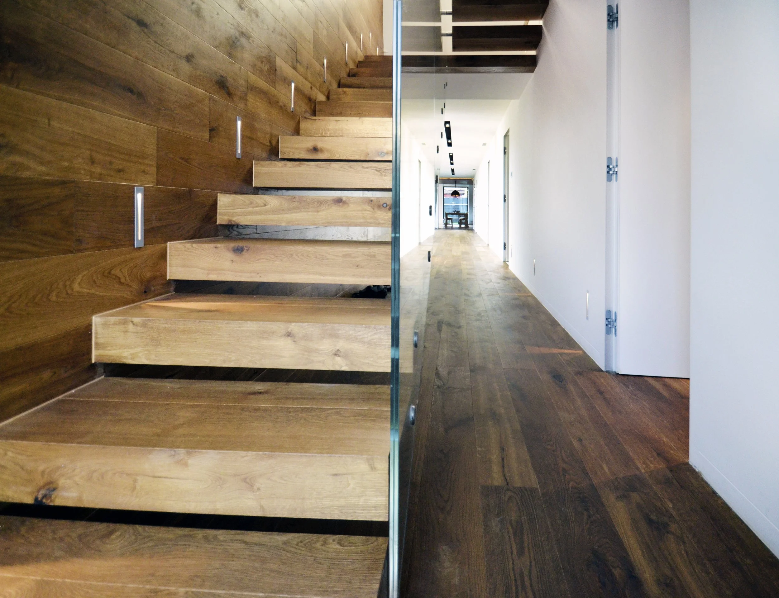 View of a modern interior space with a wooden staircase on the left, a hallway with wooden floors and white walls on the right, and a glass railing separating the staircase from the hallway.