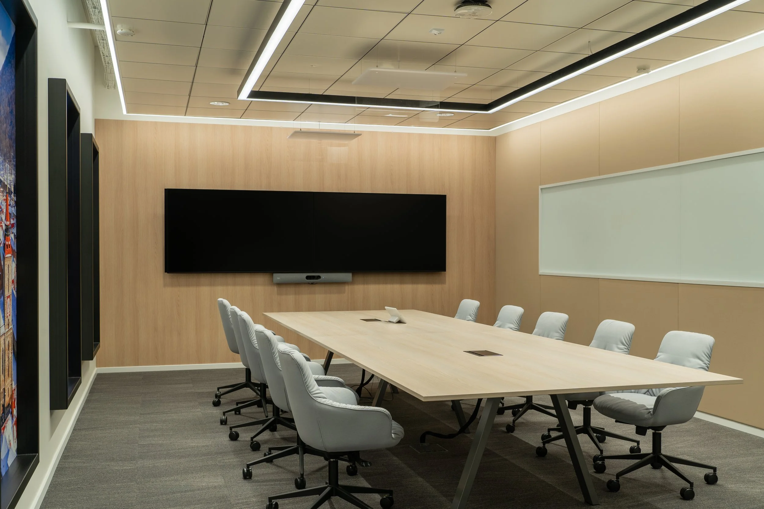 A modern conference room with a long wooden table surrounded by white cushioned swivel chairs on wheels. A large black screen is mounted on a wooden-paneled wall, and there is a whiteboard on the adjacent wall. The room has a ceiling with built-in lighting and a beige carpeted floor.