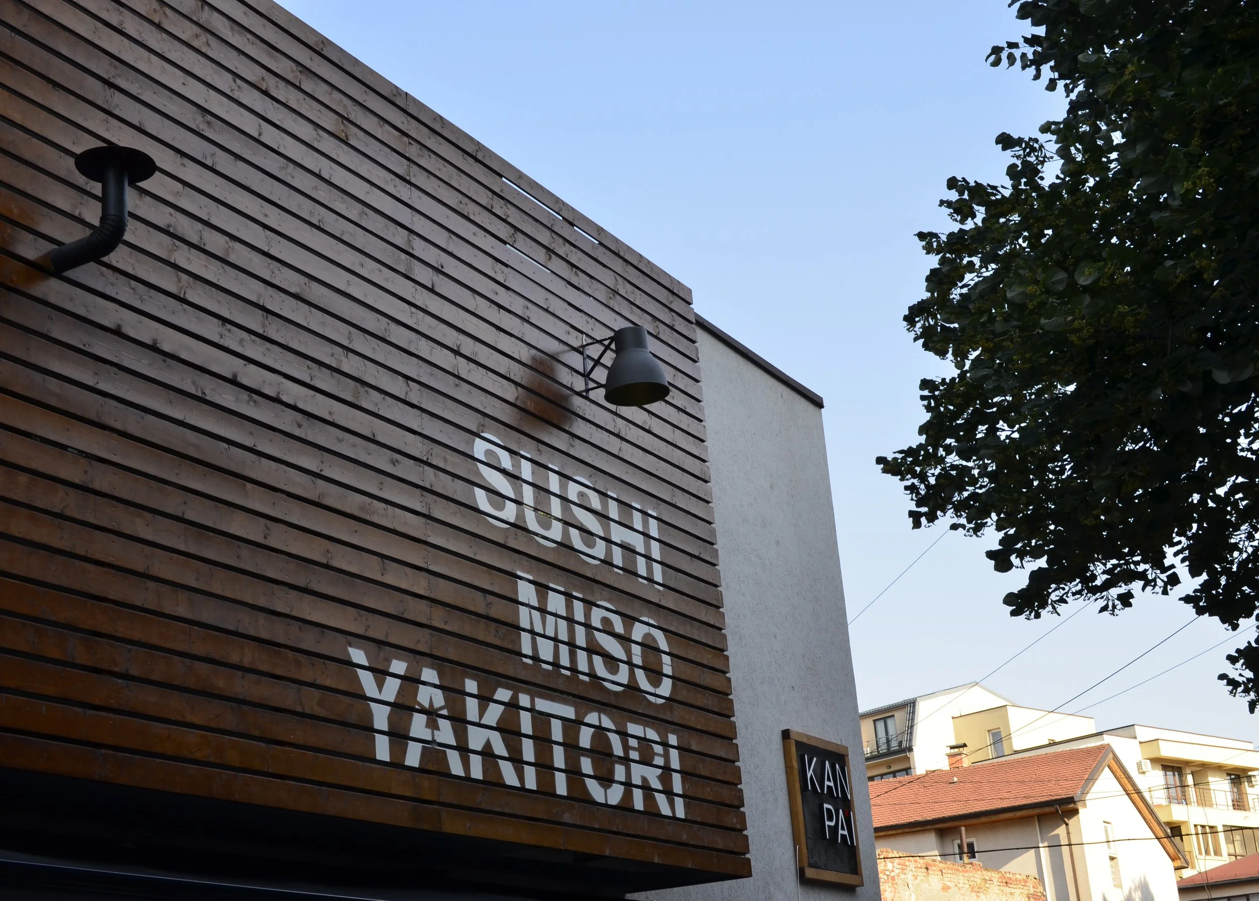 Exterior of a building with a wooden panel wall displaying the words 'SUSHI MISO YAKITORI' in white. There is a small black sign that says 'KAN PA' on the side. The sky is clear and blue, with a tree and other buildings visible in the background.