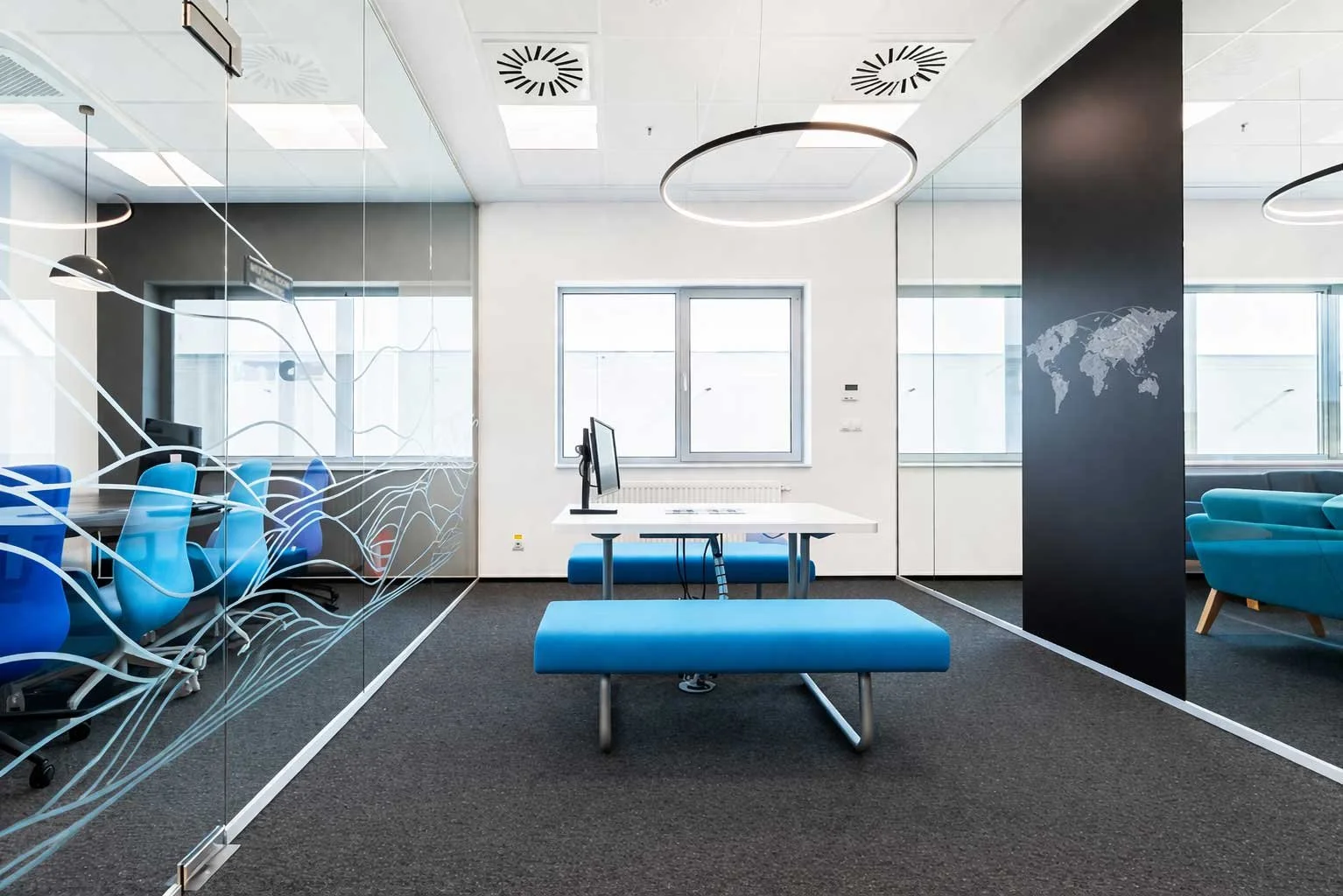 Modern office with glass walls, a black column with a world map, blue chairs, a white table, and large windows letting in natural light.