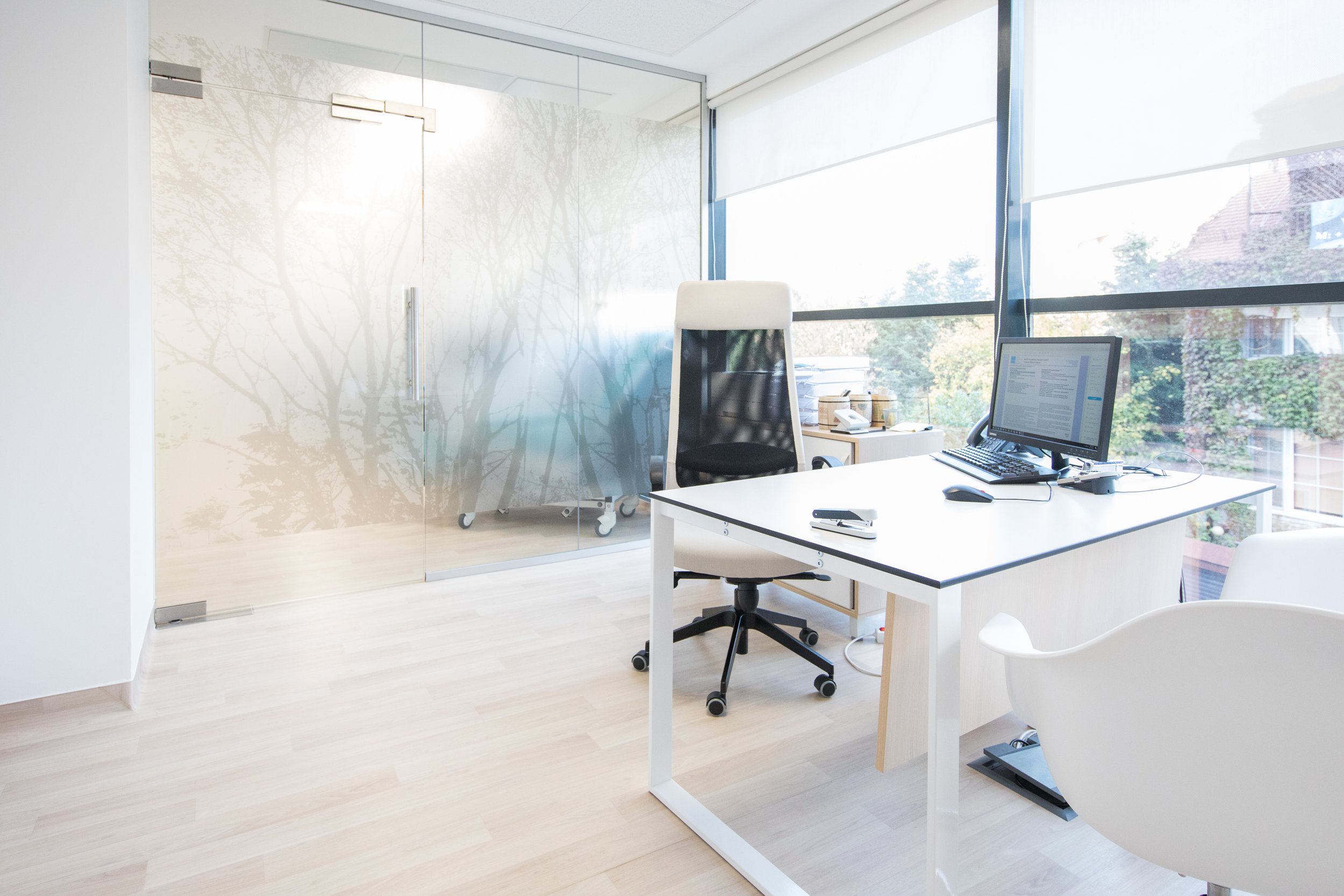 Modern, minimalistic office with a white desk, computer monitor, and black office chair near large windows with blinds. An additional white chair is visible, and a glass-walled conference room is in the background.