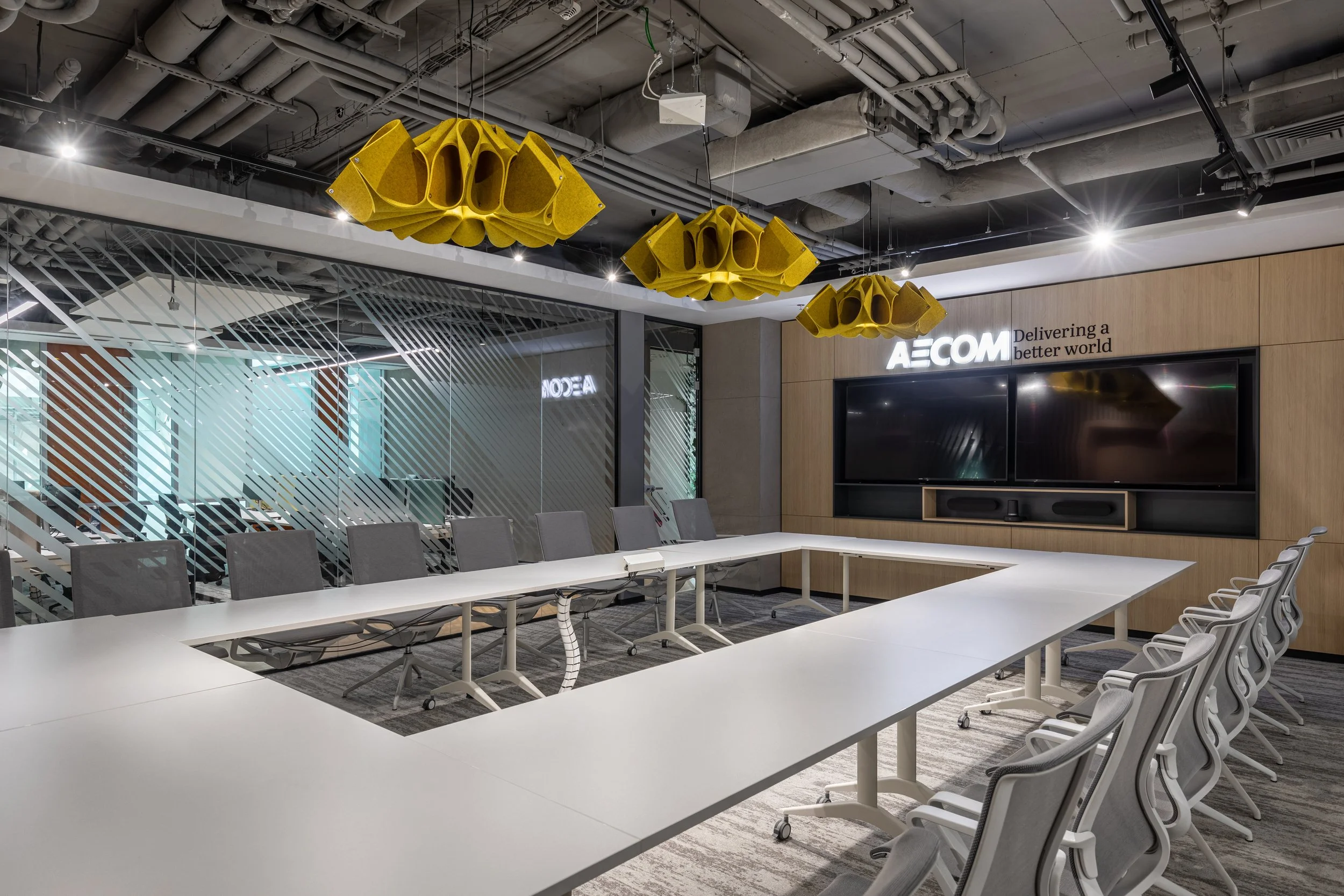 Modern conference room with a U-shaped white table, gray chairs, and yellow acoustic ceiling panels. Glass wall with diagonal stripes and company logo AECOM, tagline 'Delivering a better world' on wooden wall, large TV screen, and ceiling with exposed ductwork and lighting.