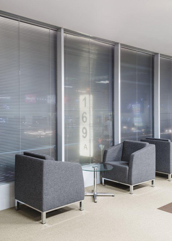 Two gray armchairs with a small round glass table between them in front of large window blinds in a waiting area or lounge.