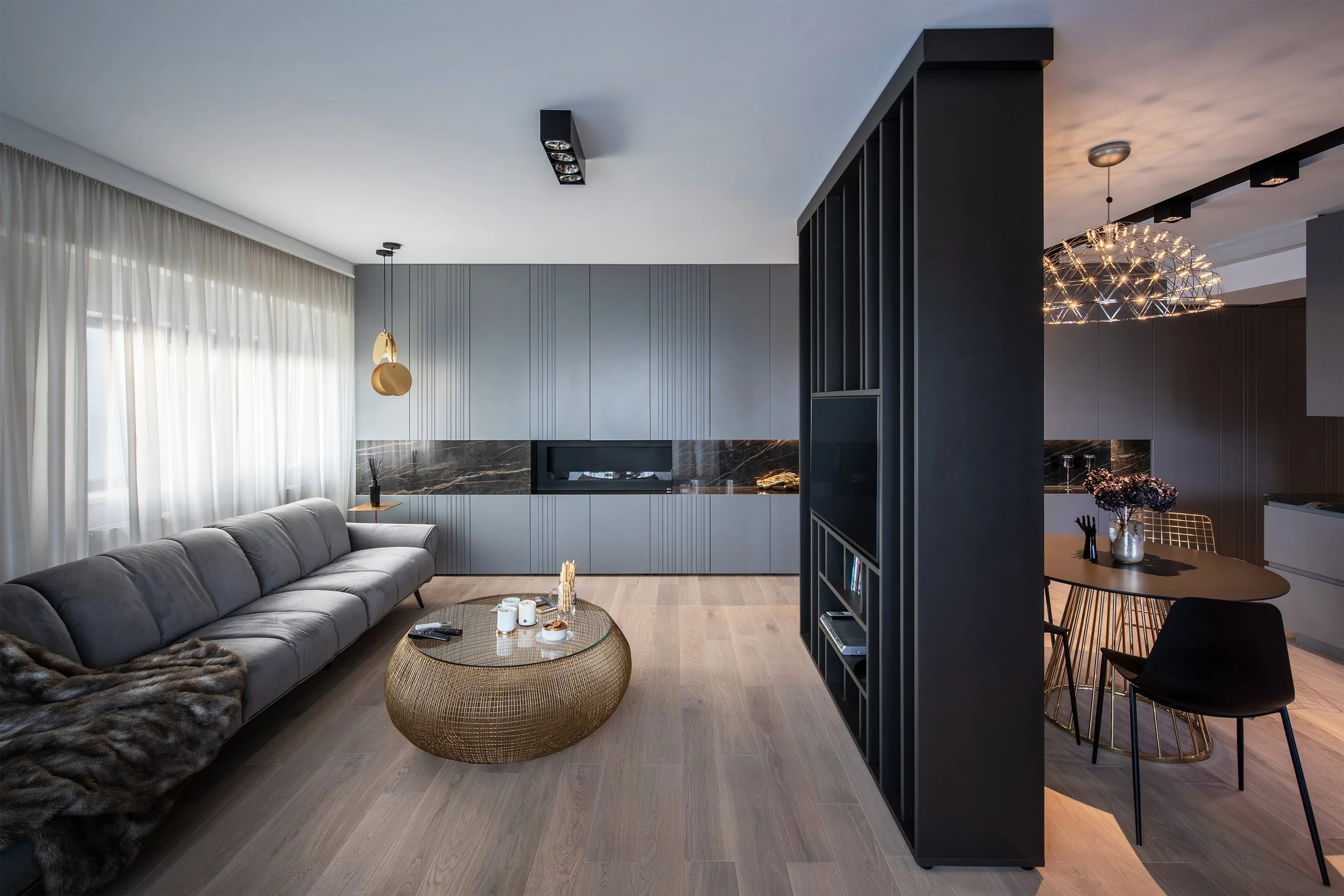 Modern living room and dining area with gray sectional sofa, round wicker coffee table, black wall-mounted TV, dark wood dining table with black chairs, contemporary chandeliers, and natural light from large window with white curtains.