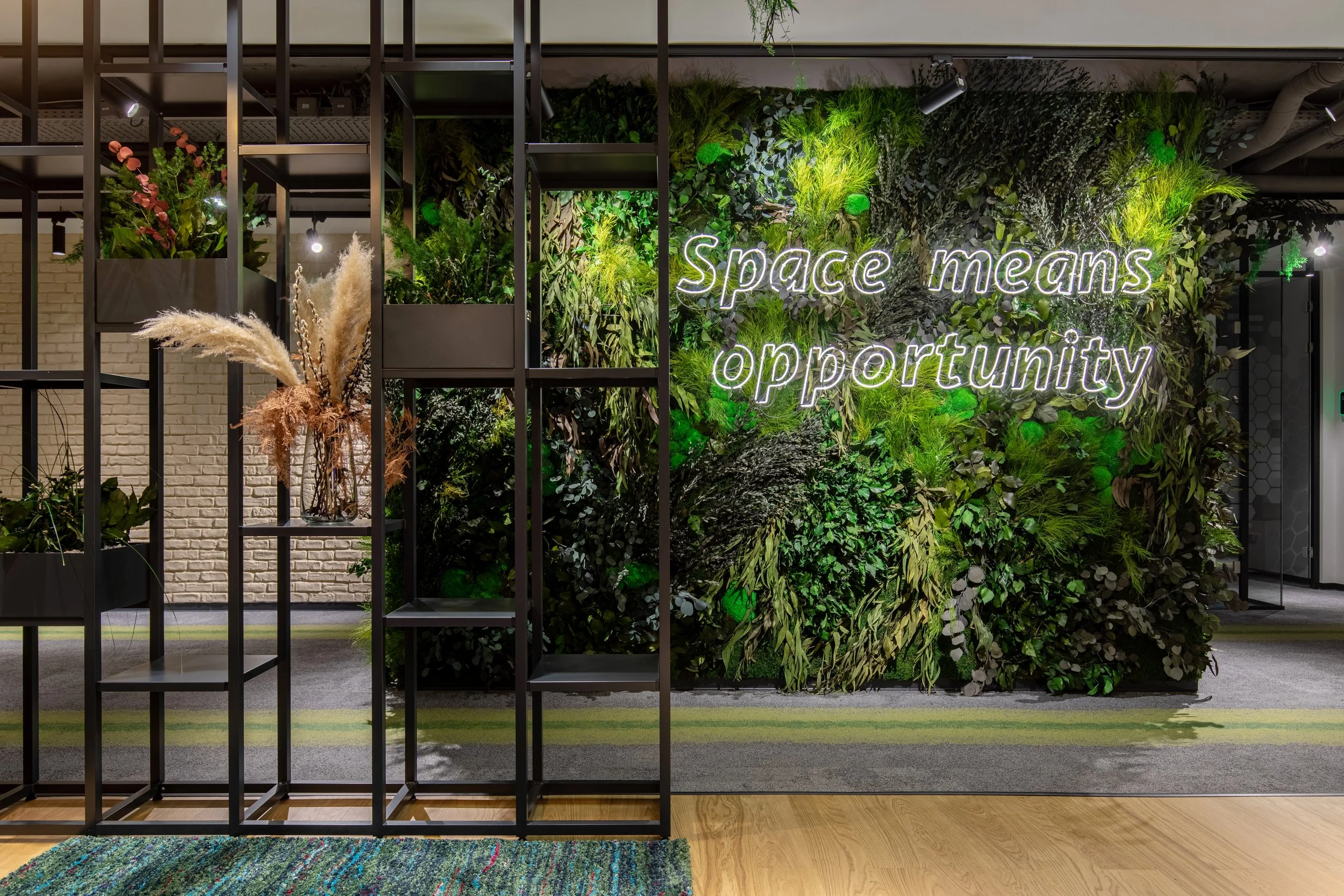 An indoor space with a green wall decorated with plants and illuminated text that says 'Space means opportunity.' There is a black metal shelf with potted plants and dried grass arrangements in front of the wall.
