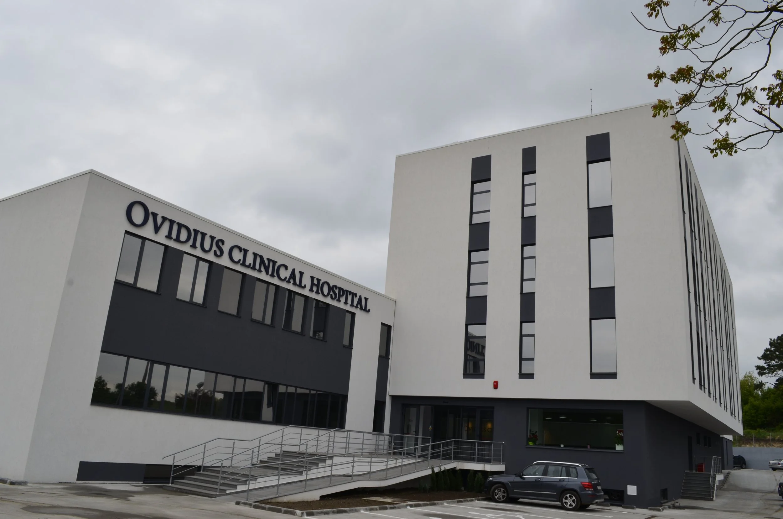 Modern hospital building with the sign 'Ovidius Clinical Hospital' on the front, featuring black and white exterior with multiple windows, parking lot in front, and cloudy sky overhead.