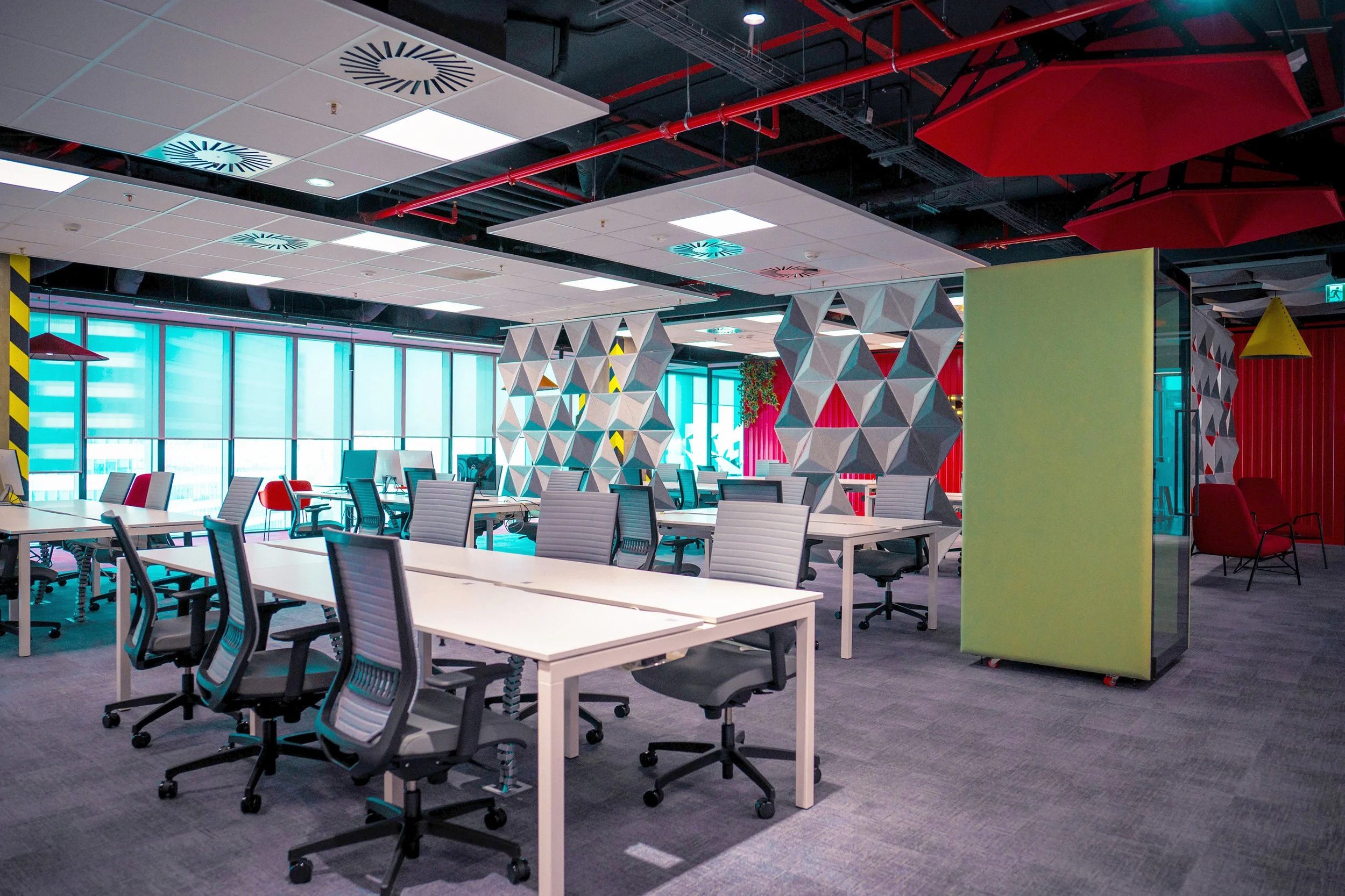 Modern open-plan office with white desks, ergonomic chairs, and geometric wall partitions, illuminated by large windows and ceiling lights.