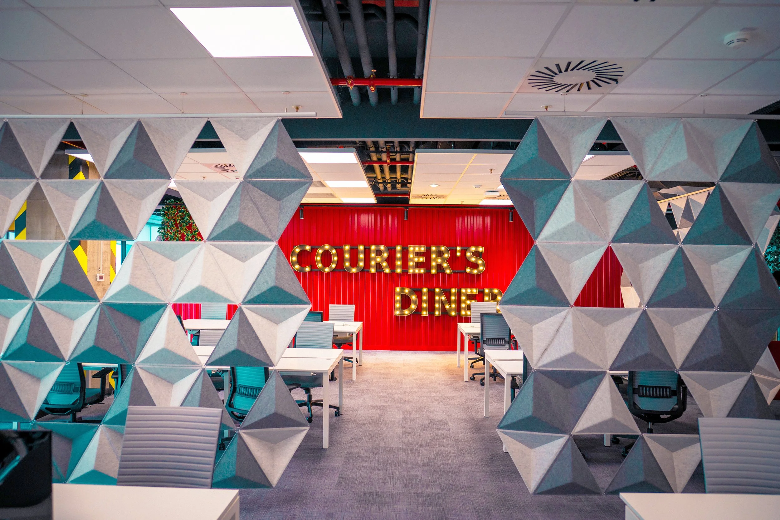 Interior of Couriers Diner with geometric decorative wall and red backdrop with illuminated sign reading 'Courier's Diner' and modern white tables and chairs.