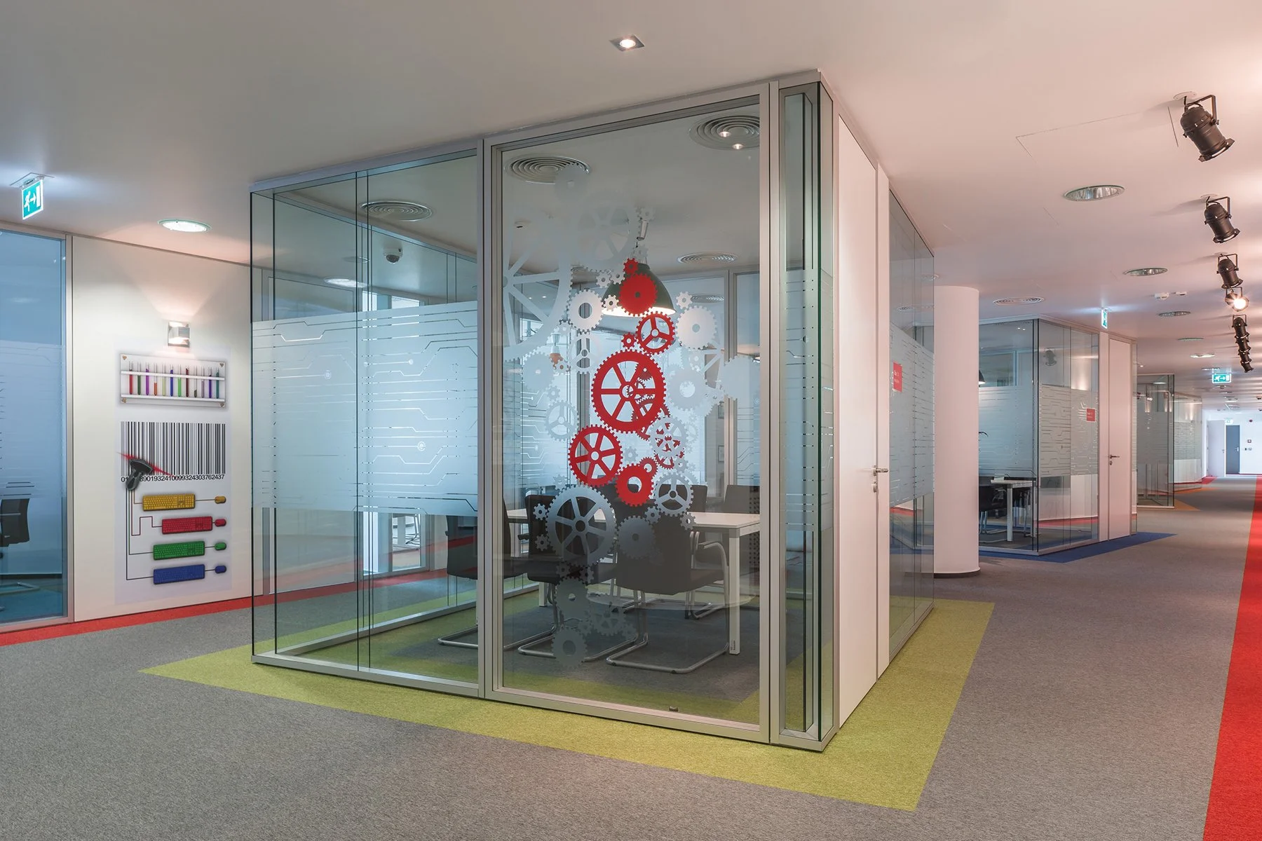 Modern office hallway with glass-enclosed meeting room decorated with red and white gears on the glass. The hallway has colorful carpet tiles and ceiling track lighting.