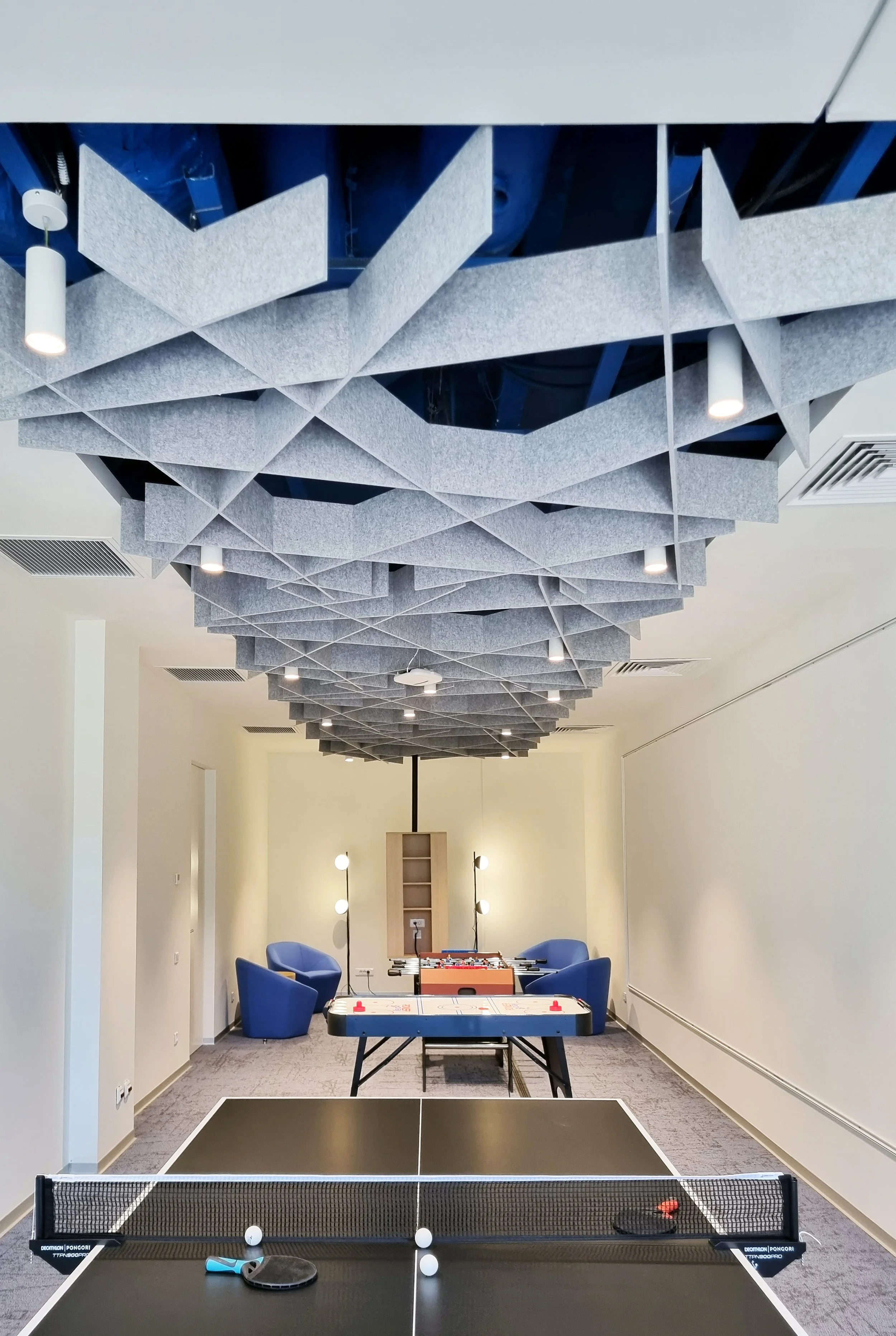 A room with a ceiling covered in acoustic panels, a table tennis setup in the foreground, a foosball table and cozy chairs at the back, and modern lighting fixtures.