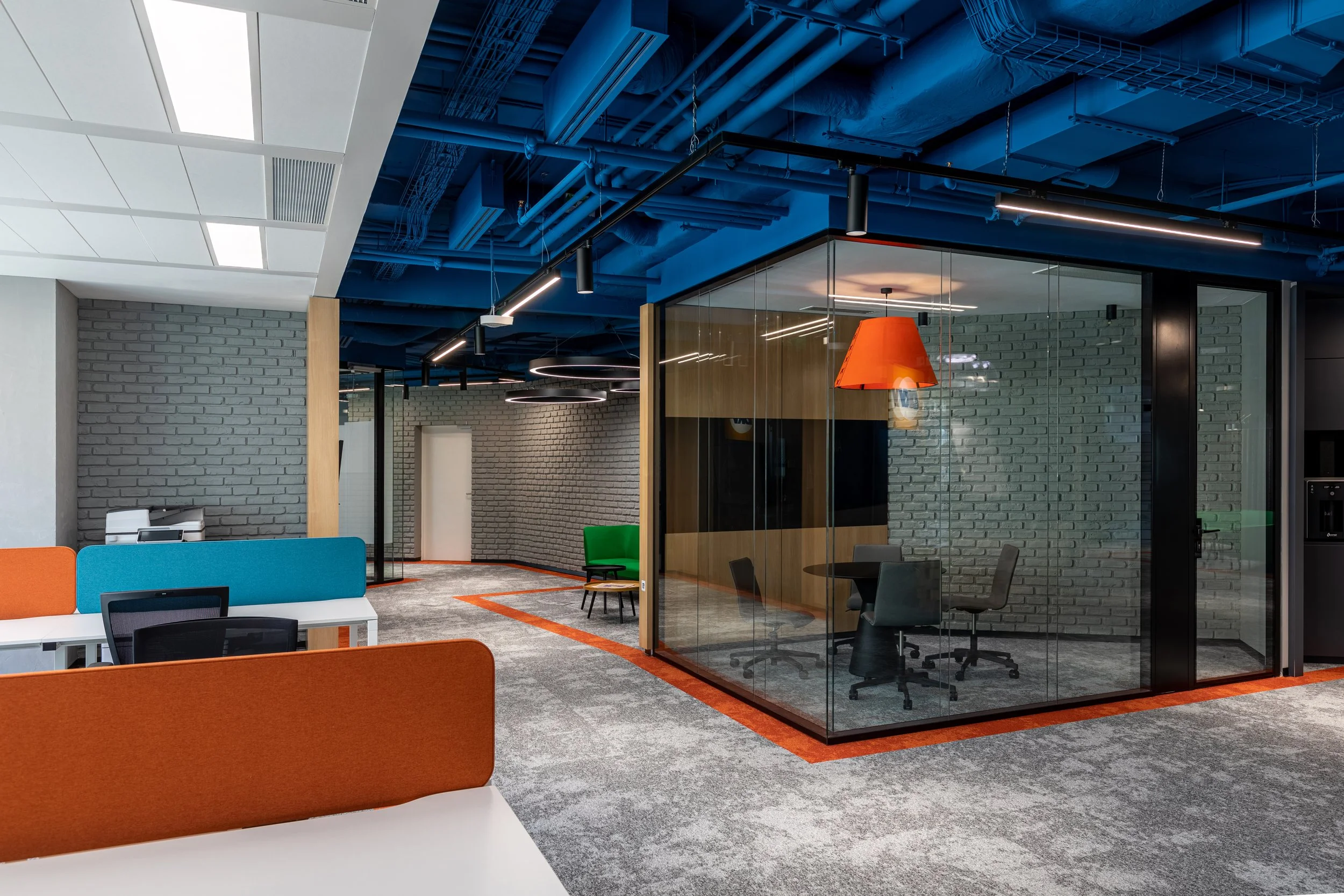 Modern office with colorful partitions, glass-walled meeting room, exposed blue ceiling pipes, and contemporary lighting fixtures.