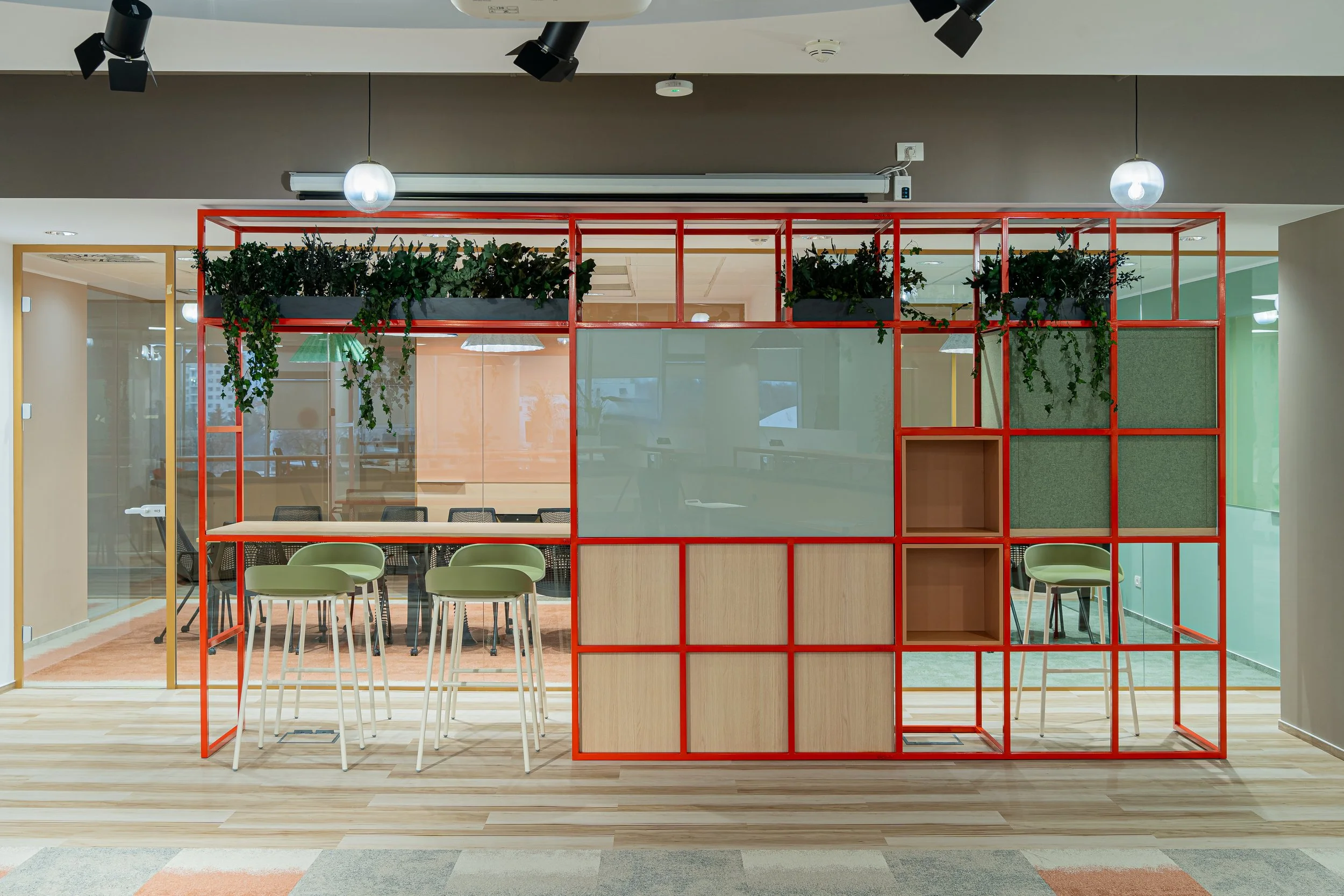 Modern office meeting room divided by a red grid partition with plants on top, glass walls, chairs, and tables inside and outside.