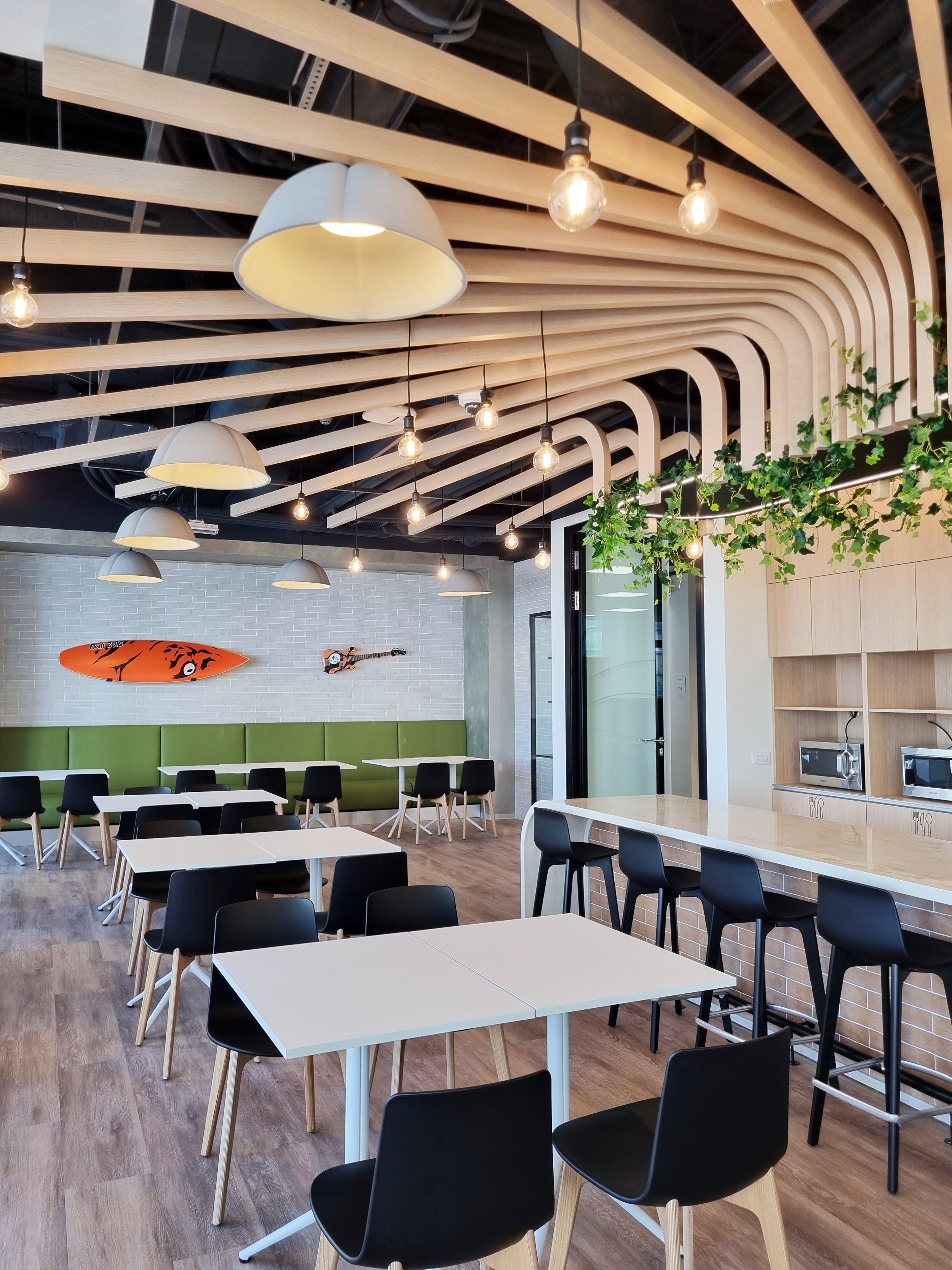 Modern cafe interior with white tables, black chairs, a green cushioned bench, decorative surfboard and guitar on a brick wall, hanging pendant lights, and wooden ceiling beams.