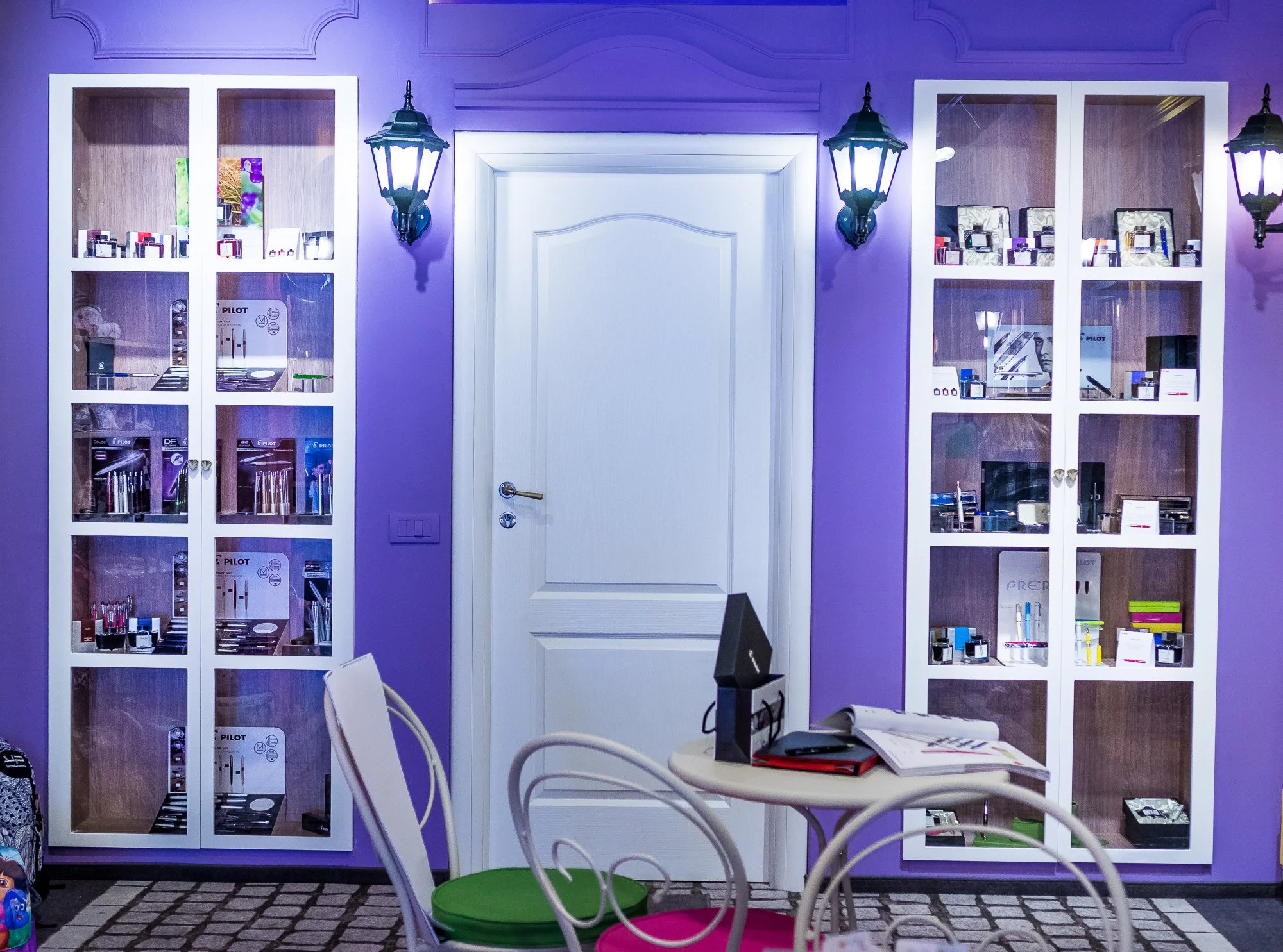 Interior of a room with purple walls, white display cabinets filled with small boxes and products, a white door, wall-mounted lanterns, a small round table with a computer monitor, a keyboard, papers, and chairs with green and pink cushions.