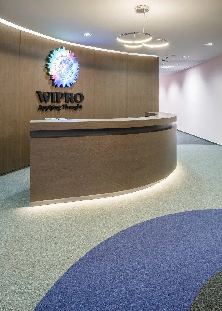 Reception area with a curved wooden desk and a illuminated WIPRO logo on the wall behind it. Modern lighting fixtures hanging from the ceiling and a carpeted floor with a circular blue pattern.