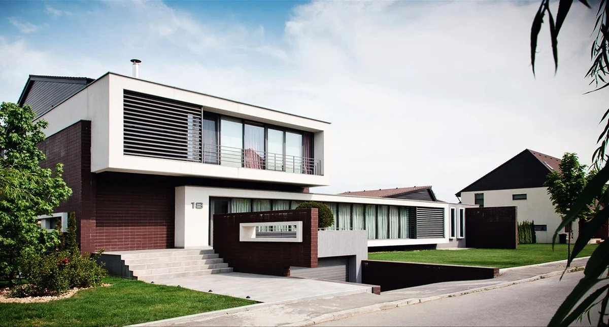 Modern two-story house with a flat roof, large glass windows, and a combination of white and dark brown exterior materials, situated on a well-maintained lawn with a sidewalk in front.