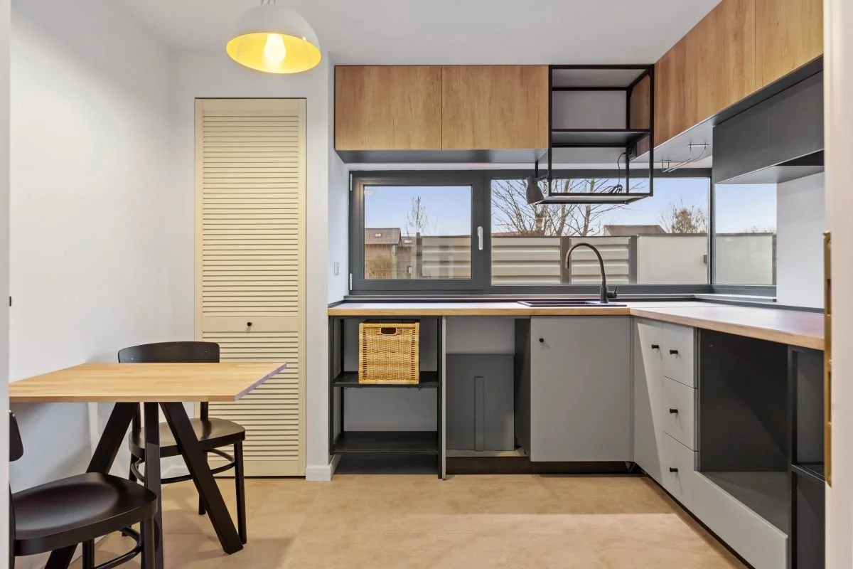 Modern kitchen with wooden cabinets, black shelving, and a large window overlooking a yard, with a small dining area featuring a wooden table and black chairs.