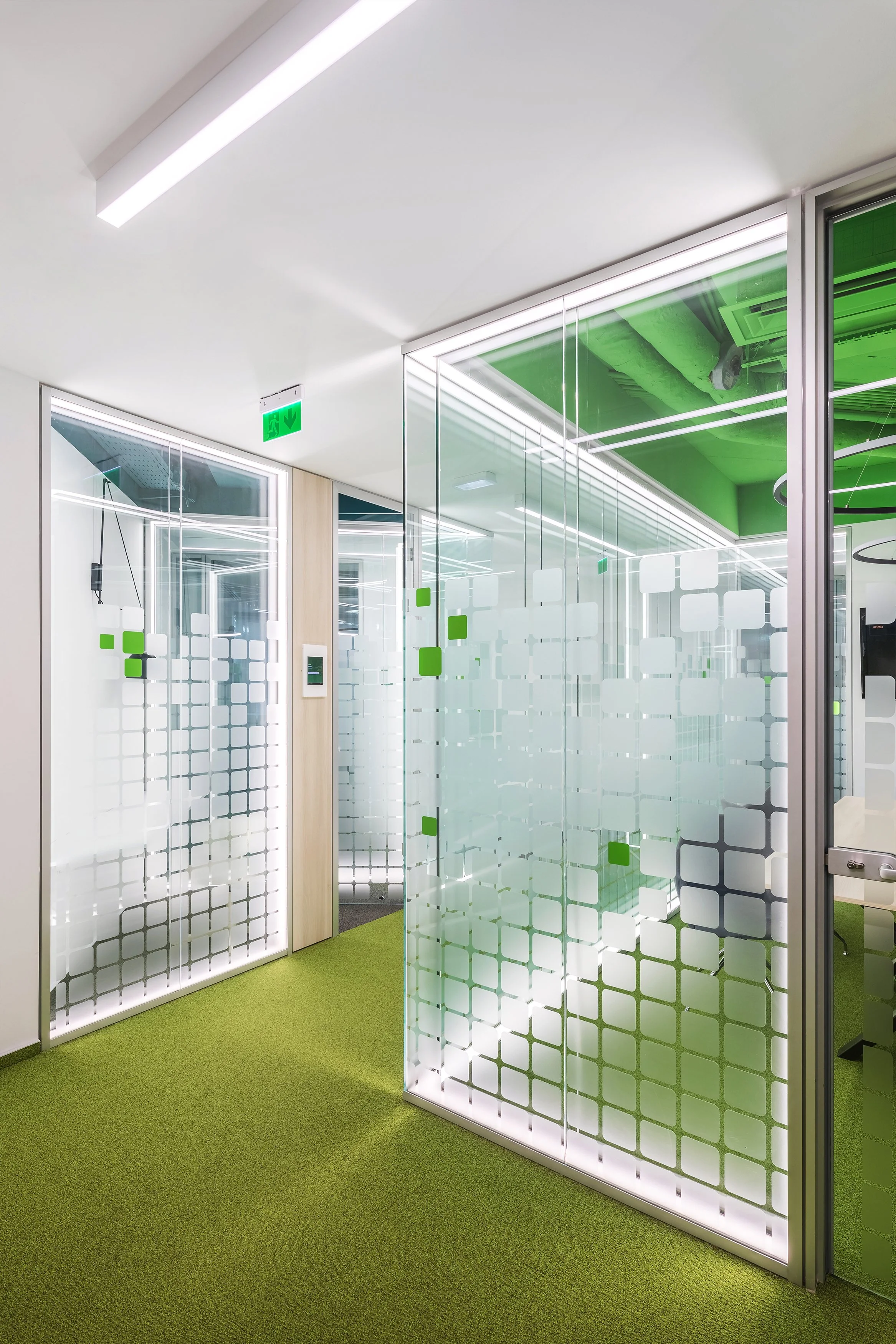 Modern office space with glass partitions, green carpet, and green ceiling accents, illuminated by bright overhead lighting.