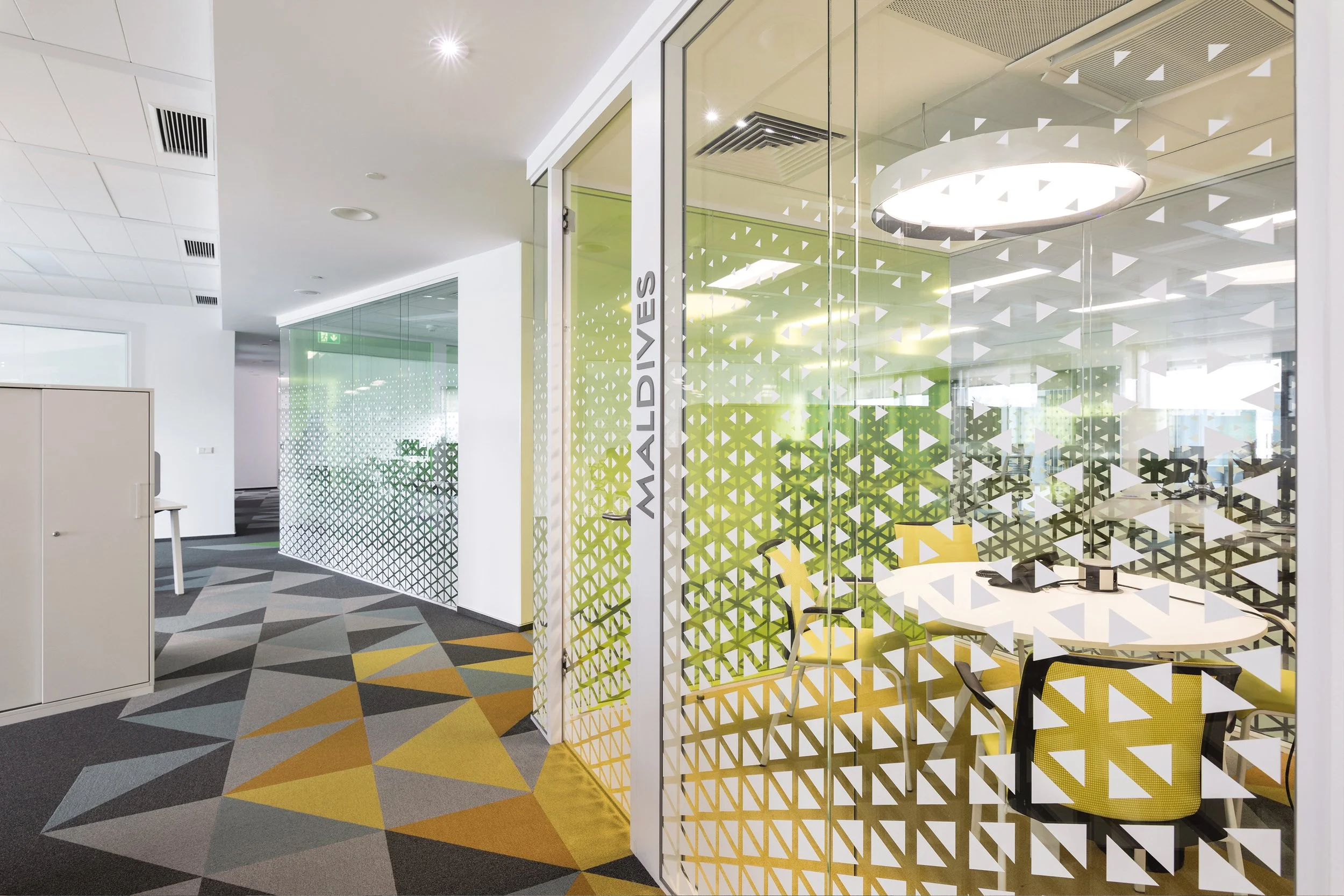 Office interior with glass partition walls decorated with geometric patterns, showing a conference room with yellow chairs and a white round table, modern lighting, and a colorful geometric patterned carpet.