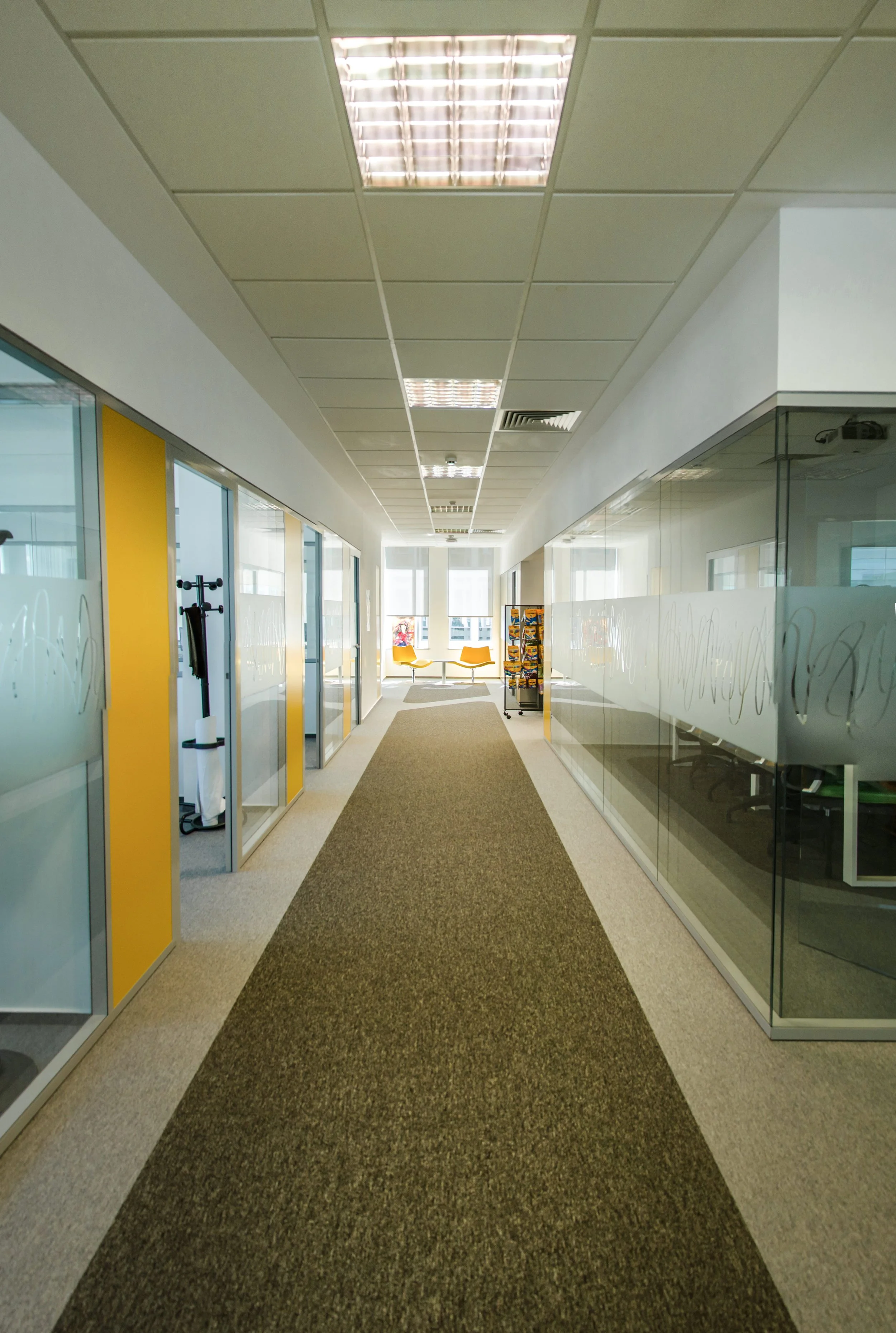 Empty office hallway with glass walls, yellow accents, and a seating area at the end with yellow chairs.