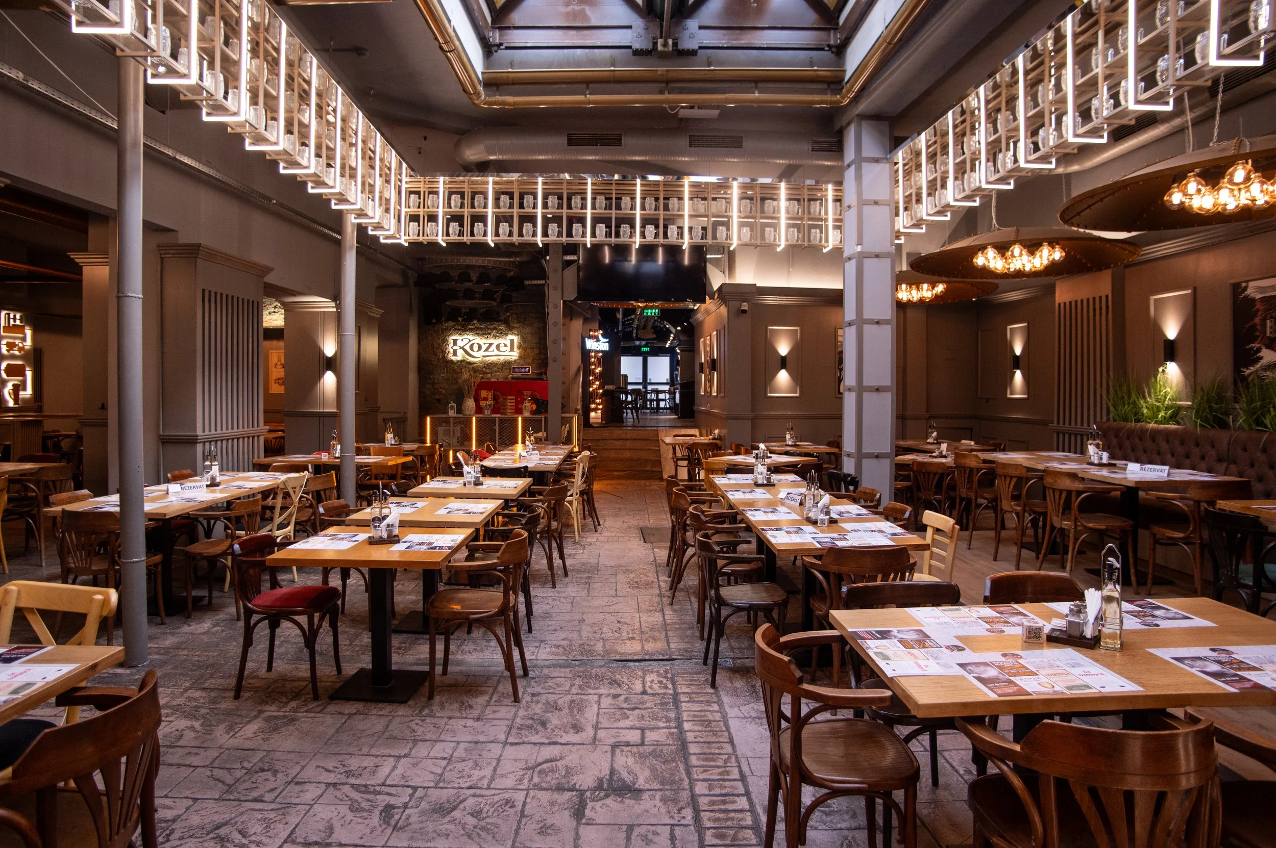 Interior of a modern restaurant with wooden tables and chairs, warm lighting, decorative wall panels, and a sign that says 'Kozel' at the back.