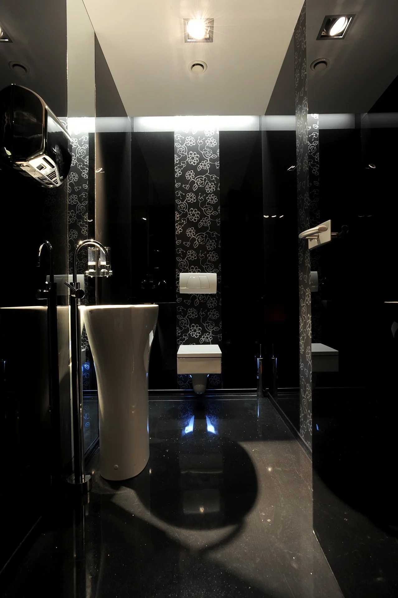 Modern black and white public restroom with a pedestal sink, wall-mounted toilet, and decorative patterned wall panels.