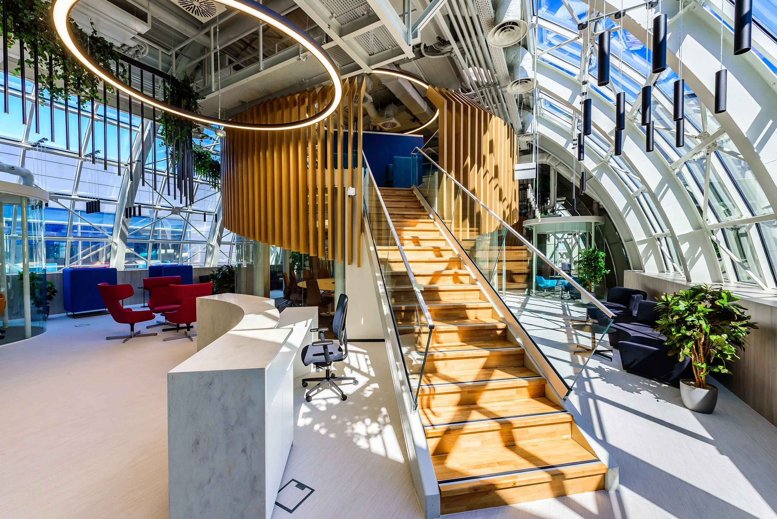 Modern office lobby with large curved glass walls, wooden staircase, seating area with red and black chairs, green plants, and contemporary lighting fixtures.