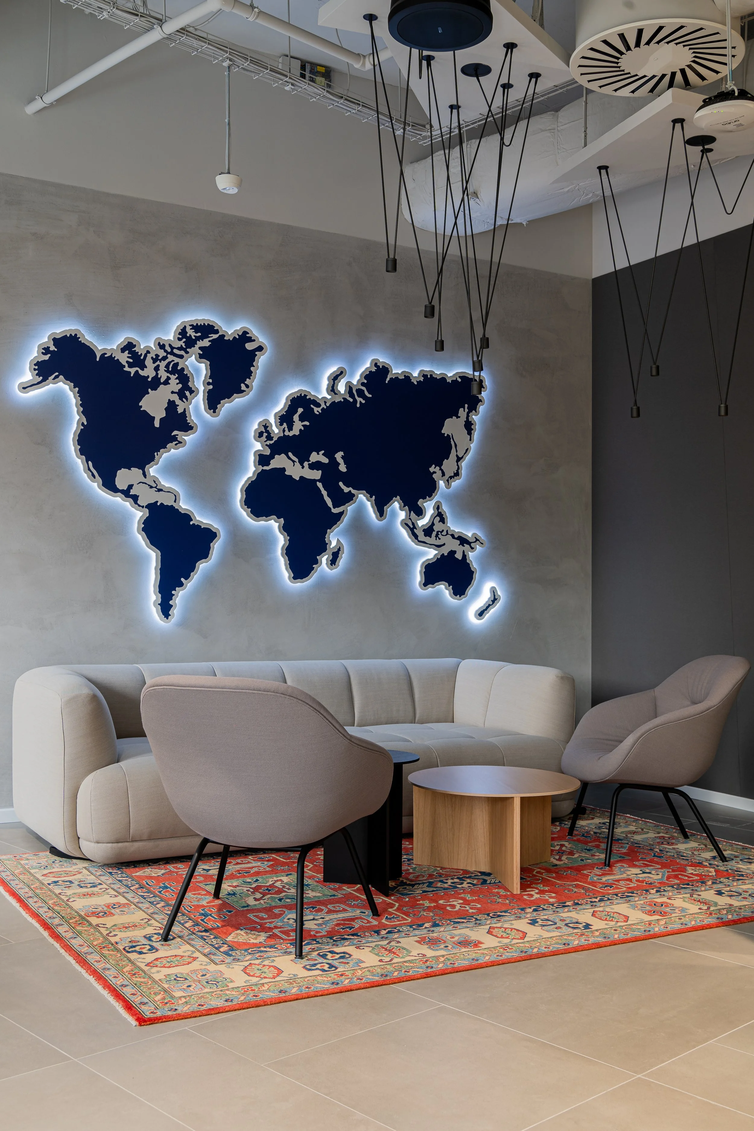 Modern office lounge area with a beige sofa, two gray armchairs, a round wooden coffee table, and a red patterned rug. On the wall behind, there is a dark blue illuminated world map decoration.
