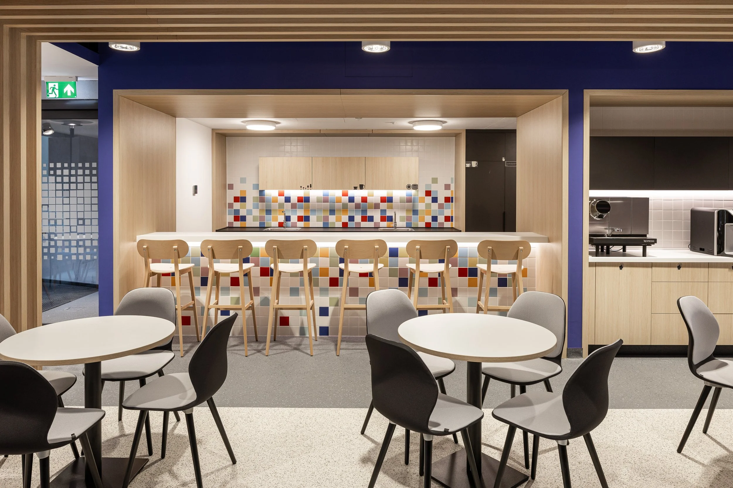 Modern cafeteria with round tables and chairs in the foreground, a bar-style counter with bar stools and colorful tile backsplash in the middle, and a kitchen area with appliances on the right, all with a contemporary design including wood accents and bold colors.