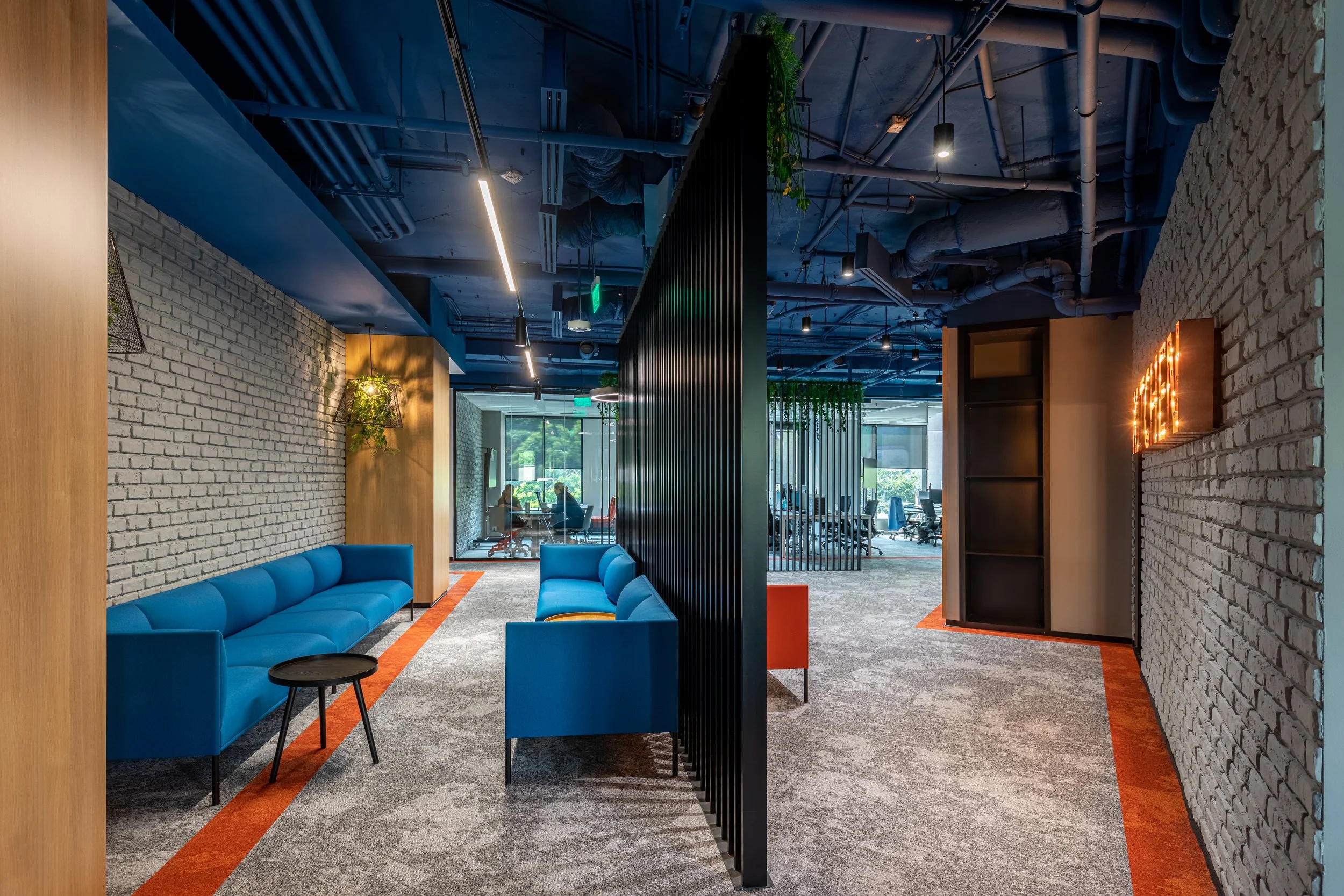 Modern office lounge with blue sofas, black side table, brick walls, hanging plants, gray carpet, and open workspace in the background.