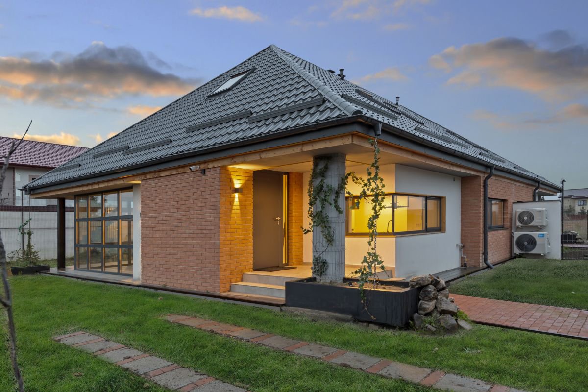 A modern house with a brick and white exterior, glass windows, and a dark gray tiled roof during sunset. The house has a small front porch with steps and an outdoor light, surrounded by a garden with grass, a stone path, and climbing plants on a concrete post.
