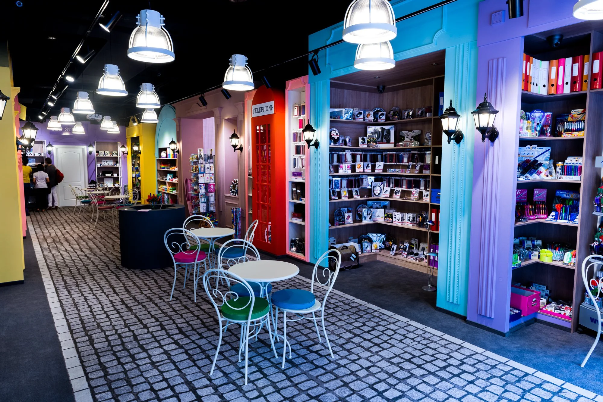 Colorful bookstore or stationery store with shelves of notebooks and pens, glass tables and chairs, decorative lighting, and a red telephone booth