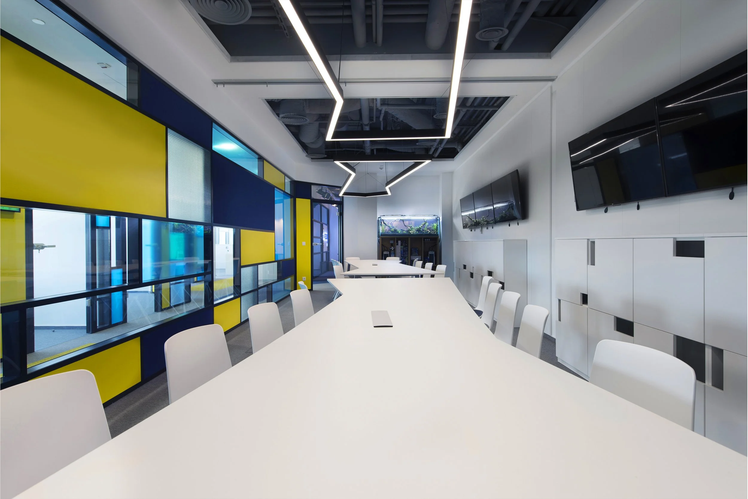 Modern conference room with a long white table, white chairs, multiple wall-mounted screens, a glass office door, and a decorative ceiling with geometric lighting fixtures.