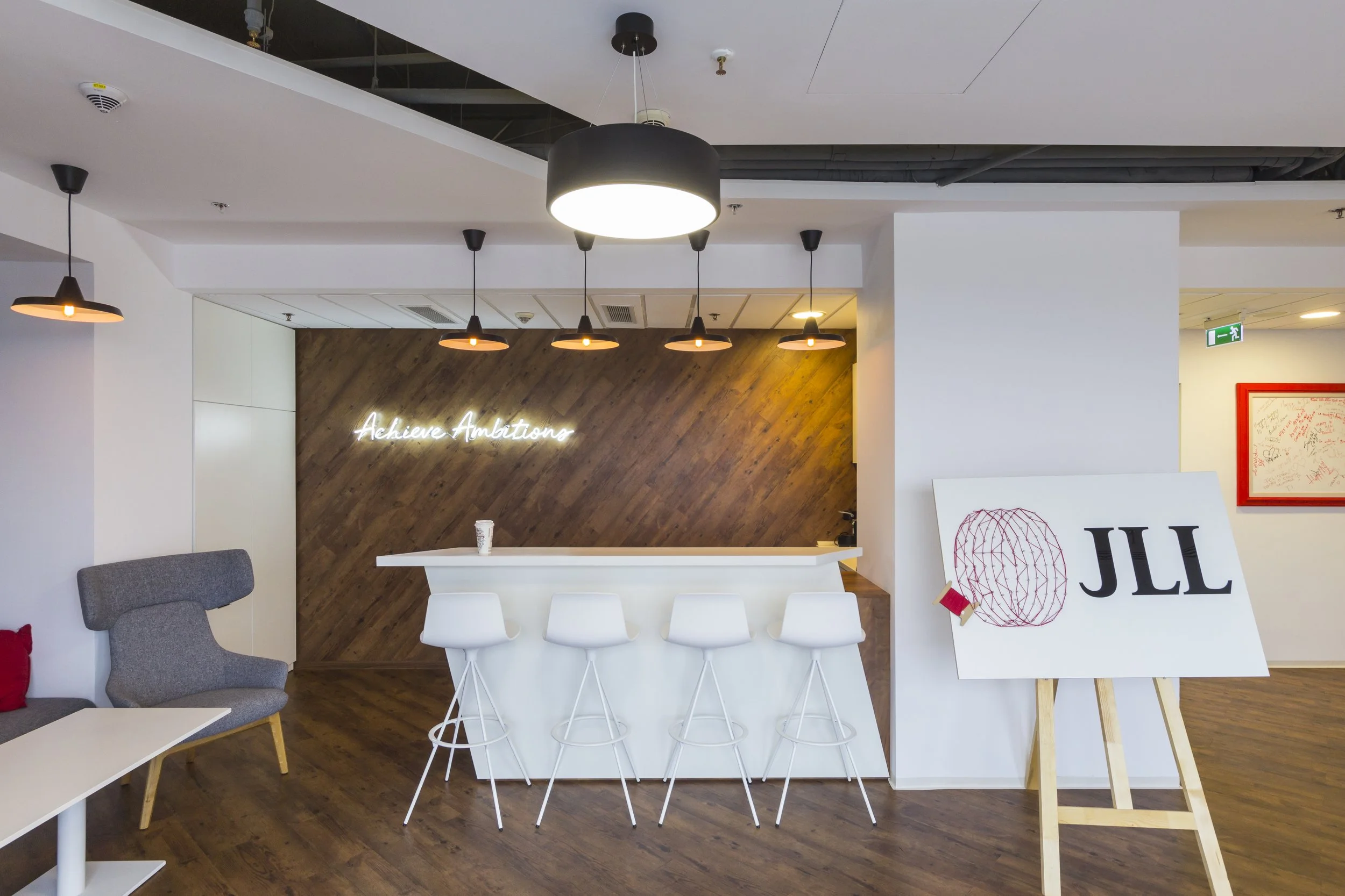 Modern office lounge with white bar stools, a high white counter, wooden wall with neon sign that reads 'Achieve Ambitions', and JLL branding on an easel