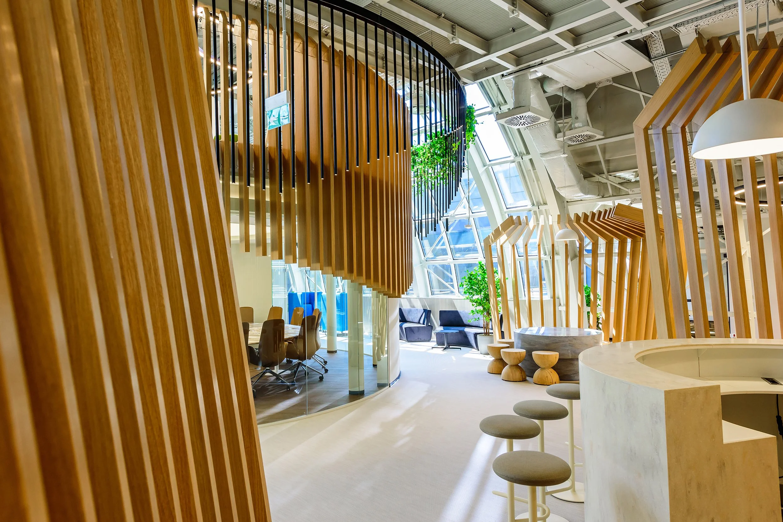 Modern office interior with wooden slats, glass walls, and ample natural light from large windows with skylights. There are various seating areas, including chairs, stools, and small tables, accented with indoor greenery.