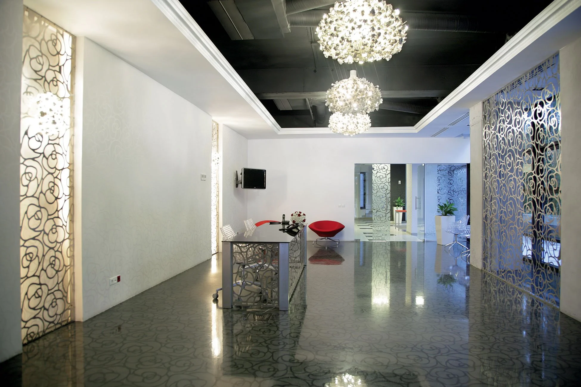 Modern hotel lobby with white walls, patterned metallic flooring, decorative wall panels, pendant chandeliers, a red lounge chair, and a desk with a computer and flowers.