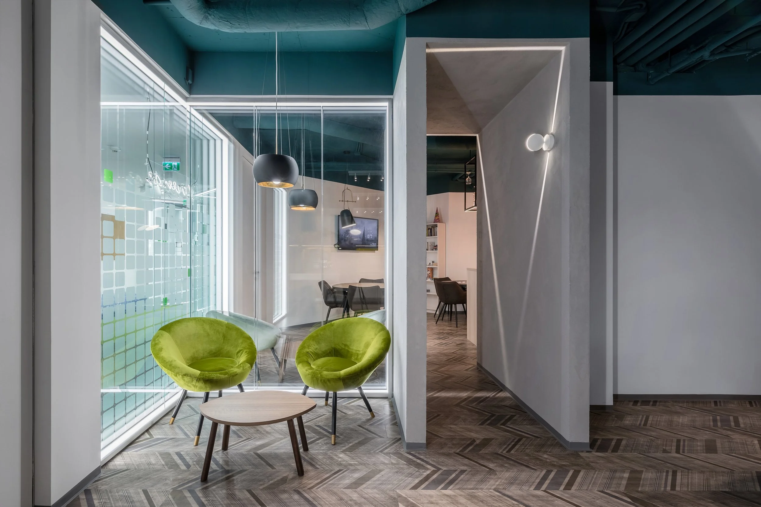 Modern interior space with two lime green velvet chairs, a small round wooden table, and pendant lights hanging from the ceiling. Glass walls with a grid design, and a hallway with dark wood flooring and wall lighting.
