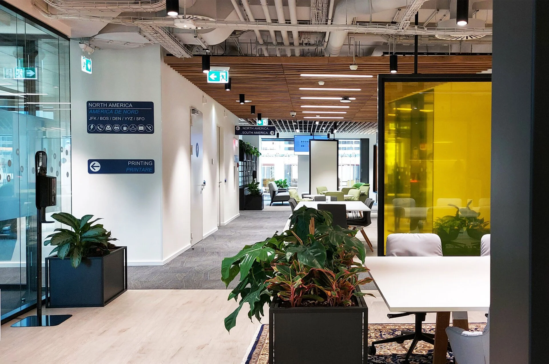 Modern airport lounge with seating area, potted plants, and signage indicating directions for North America and printing services.